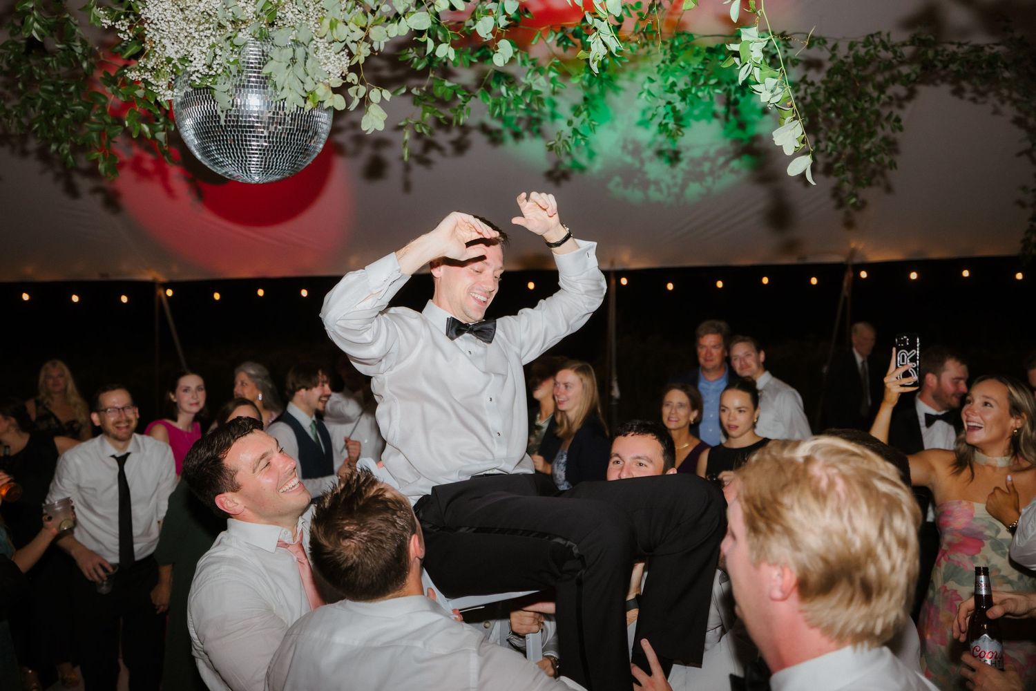 Groom in black tuxedo is hoisted up by groomsmen celebrating on the wedding reception dance floor.