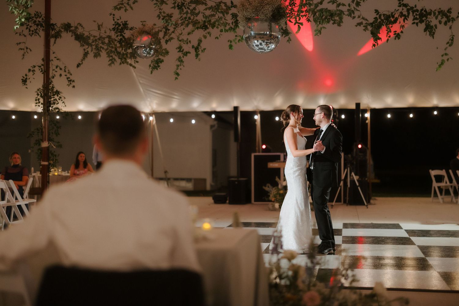 Bride and groom share their first dance on a checkered floor under romantic red lighting in a tent.