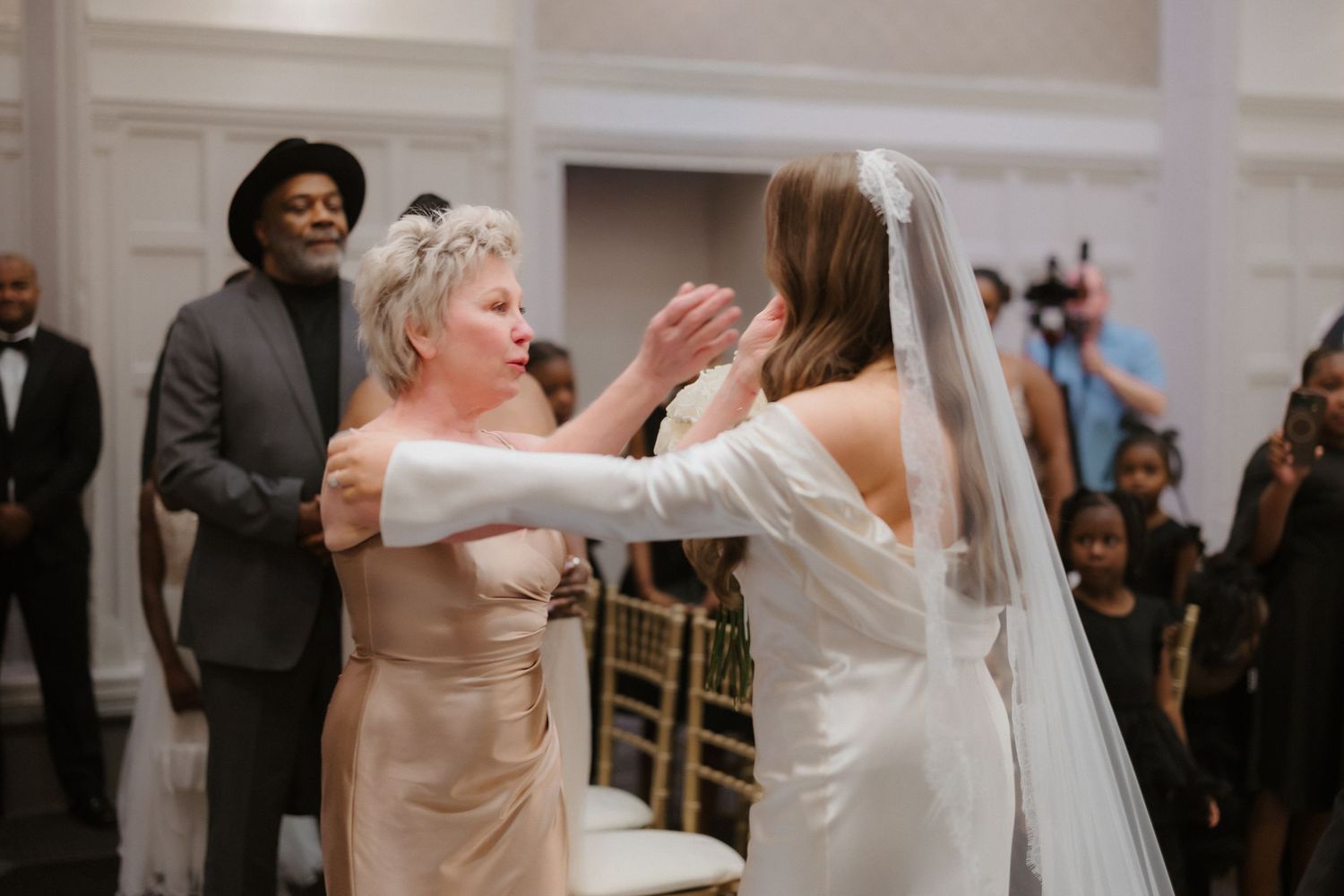 Bride in white gown and veil shares emotional moment with older woman in champagne dress at wedding reception.