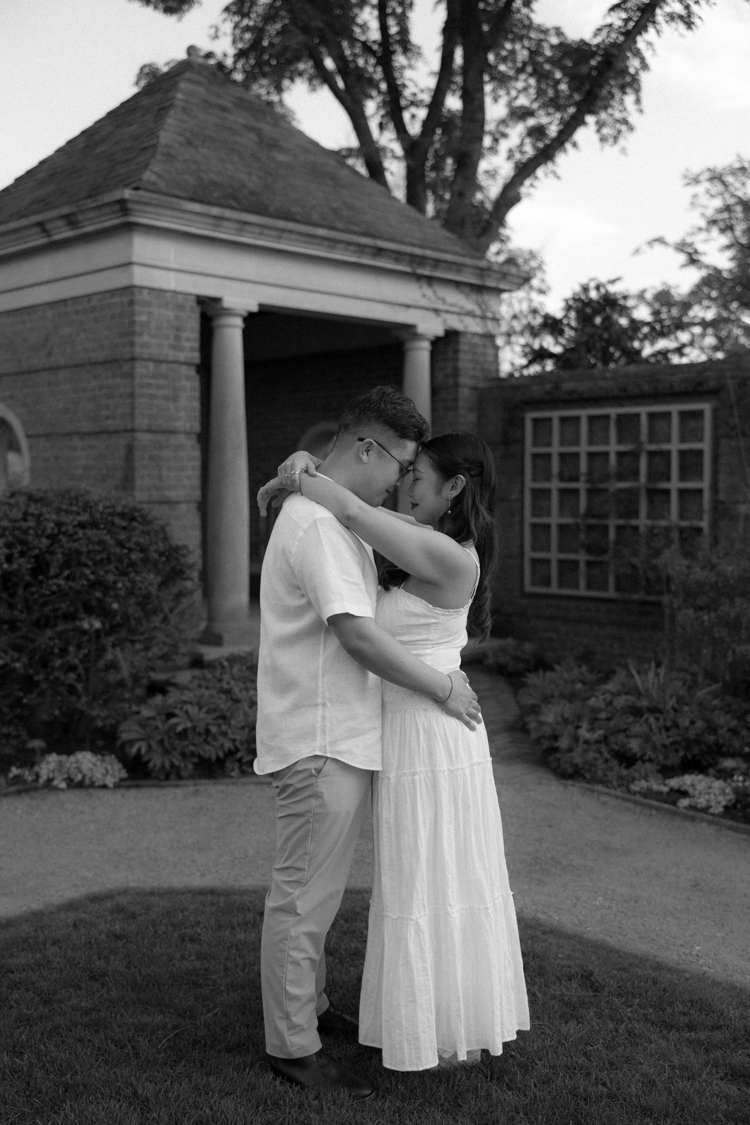 Black and white photo of a couple embracing and kissing in a garden with a stone gazebo backdrop.
