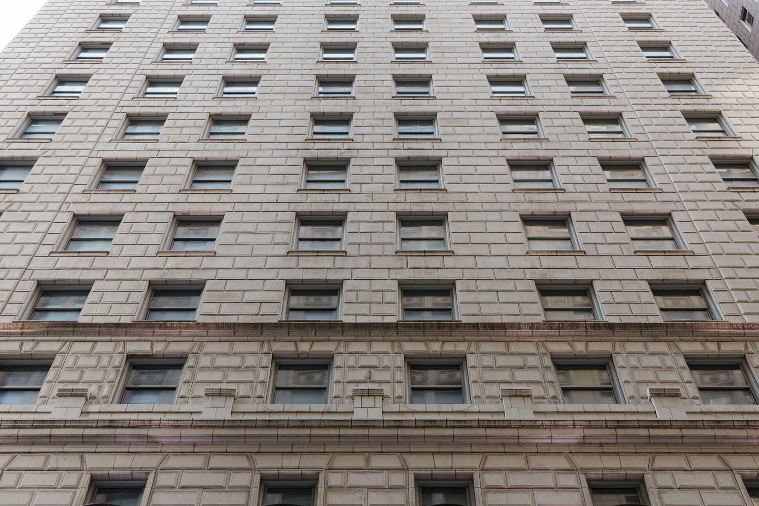 Looking up at a tall beige stone office building facade with rows of rectangular windows in an urban setting.