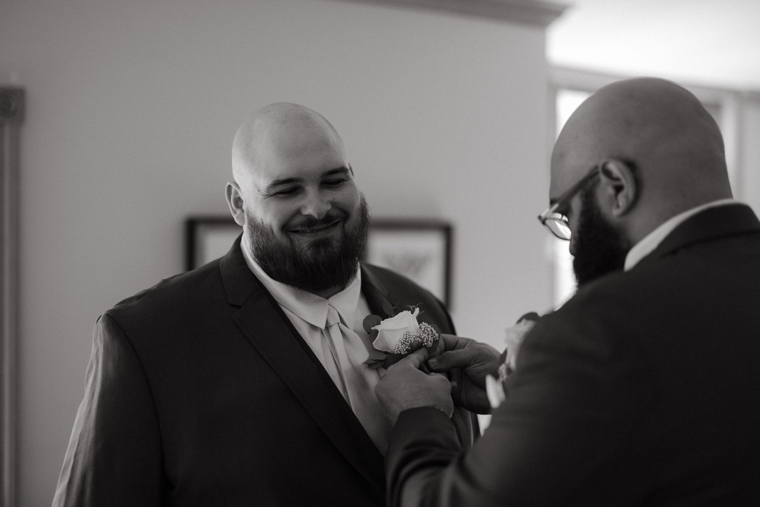 Black and white photo of a groom smiling as his groomsman pins a boutonniere on his suit lapel.