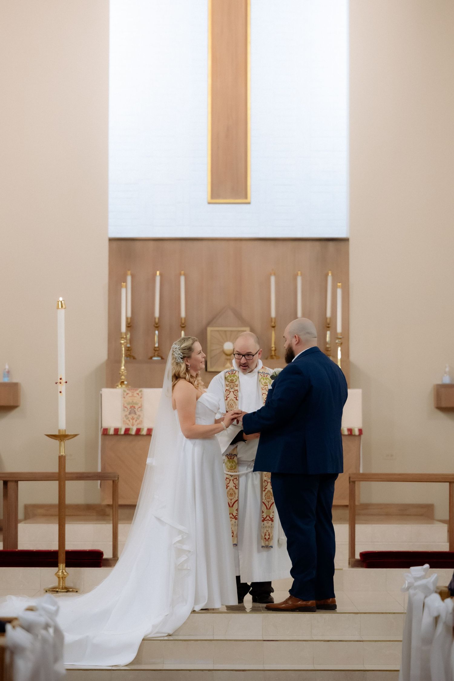 Bride and groom exchange vows at church altar with officiant, candles, and wooden cross backdrop.
