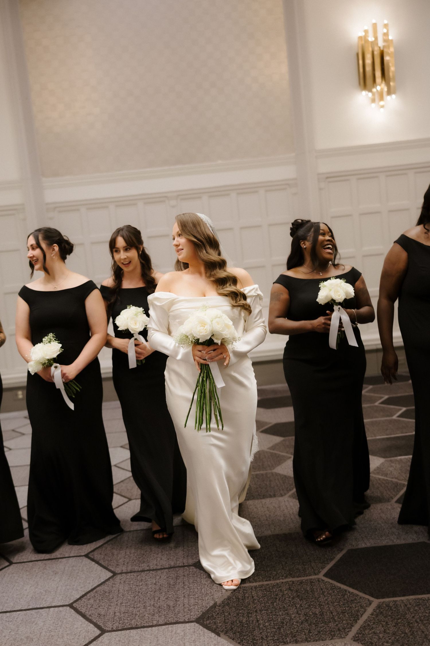 Bride in white off-shoulder gown walks with bridesmaids in black dresses, all holding white bouquets in elegant venue.