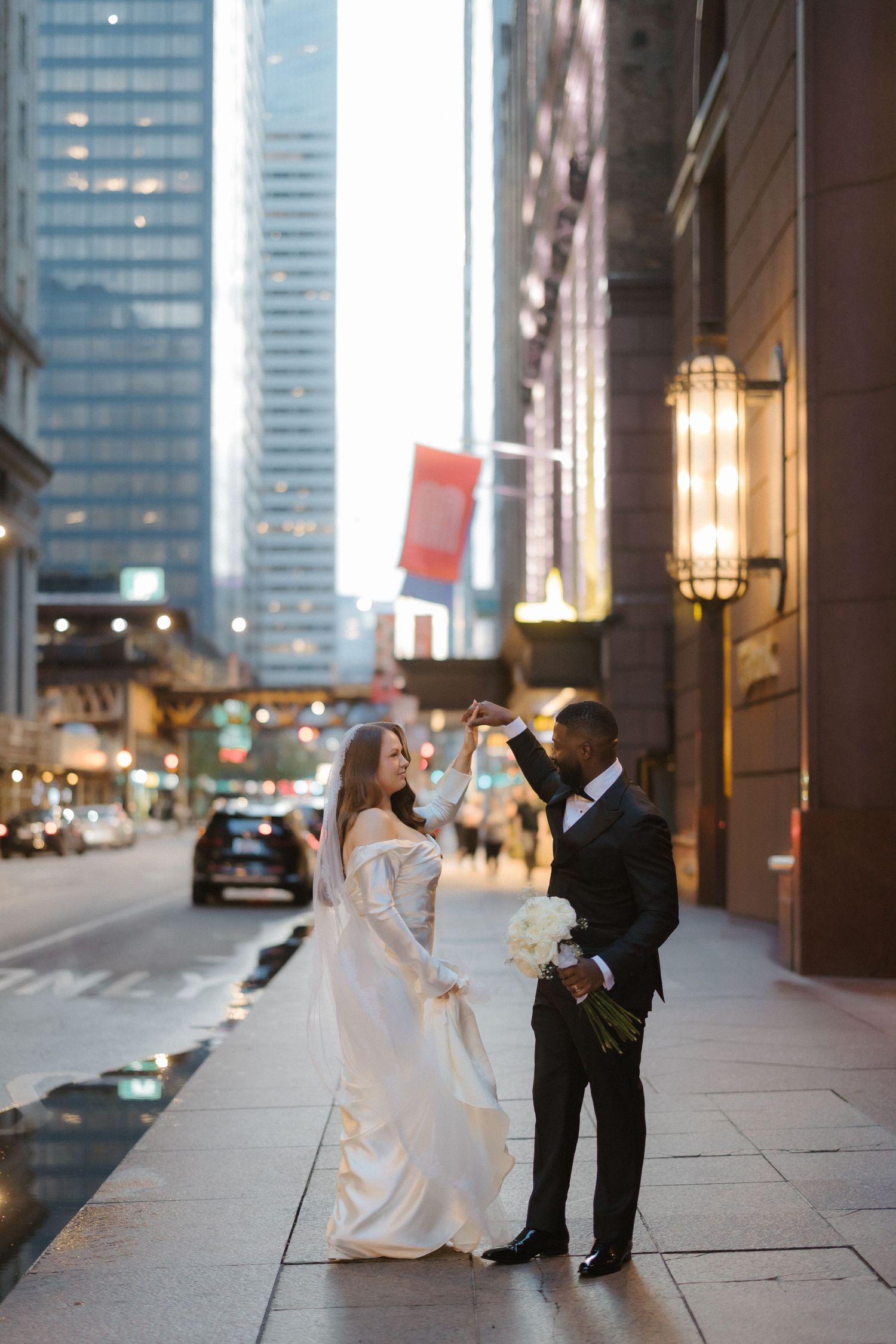 Interracial couple dancing on urban sidewalk, bride in white gown holding bouquet, groom in tuxedo, city skyscrapers backdrop.