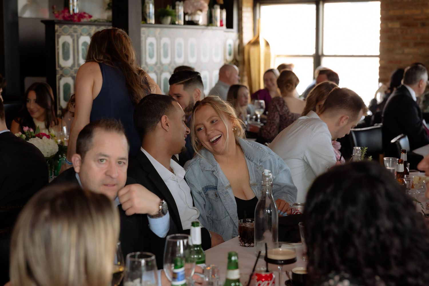Guests laughing and socializing at a lively indoor dinner event with tables set with glasses and bottles.