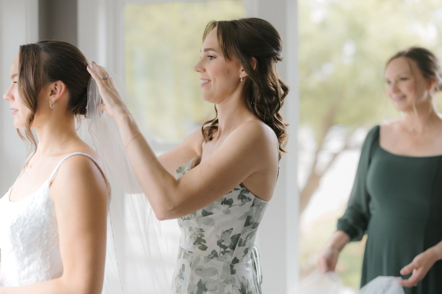 Bridesmaid in floral dress places veil on bride in white lace gown as woman in green dress watches.