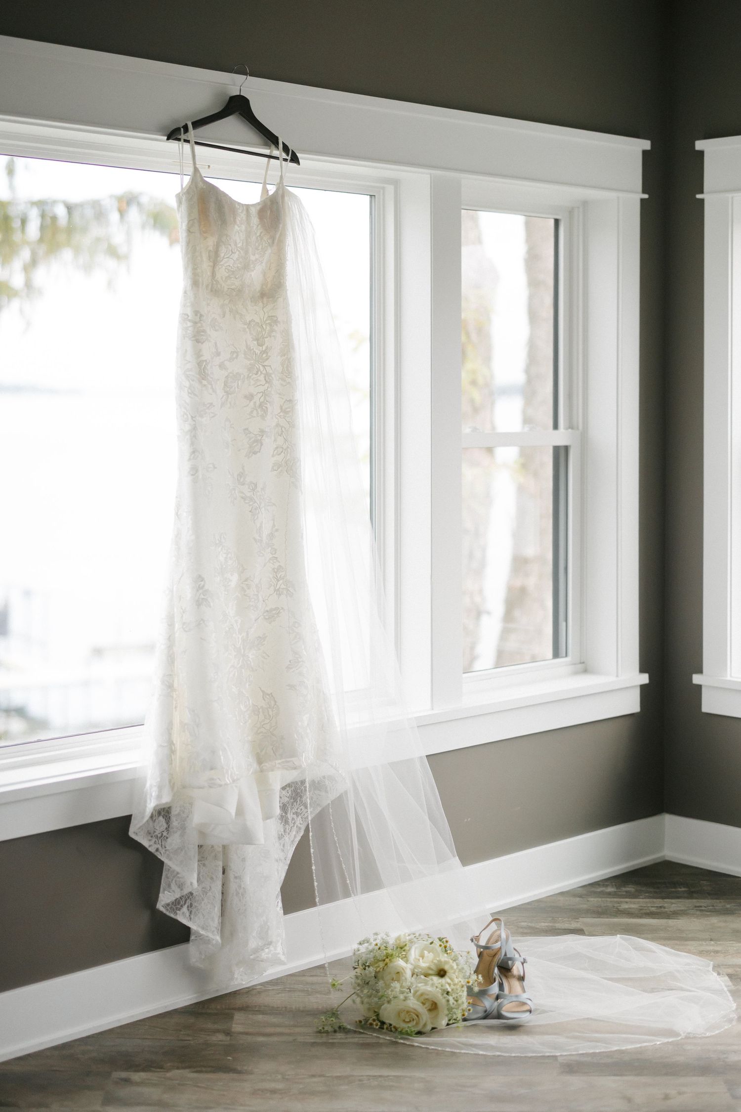 White lace wedding gown hanging in a bright window with bridal bouquet and shoes below.