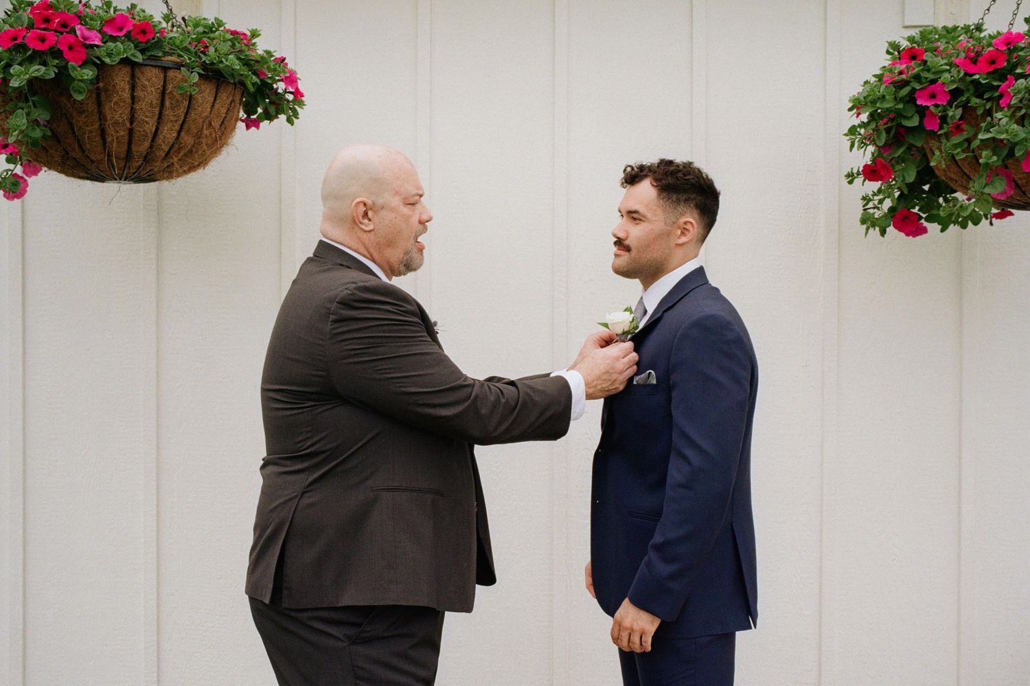 A man in a gray suit pins a boutonniere on a groom in a navy blue suit at a wedding ceremony.