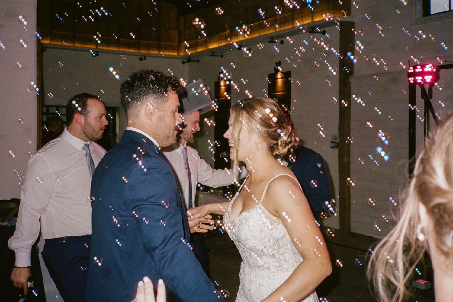 Bride and groom dance joyfully surrounded by bubbles at their wedding reception, groom in blue suit.