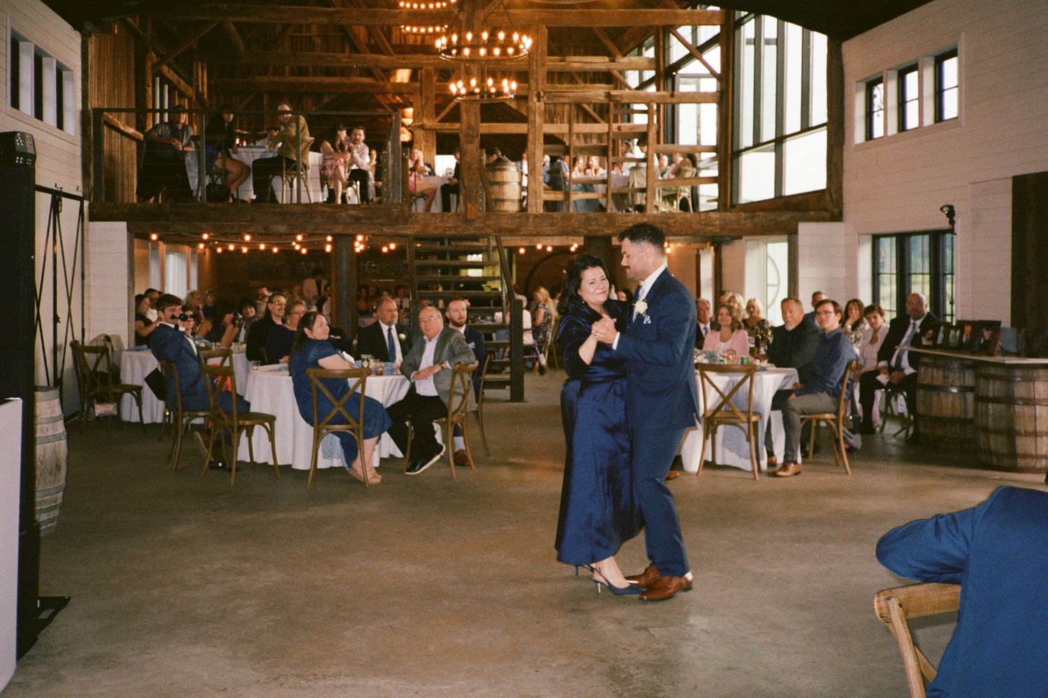 Couple shares first dance in rustic barn wedding venue with string lights and seated guests watching.