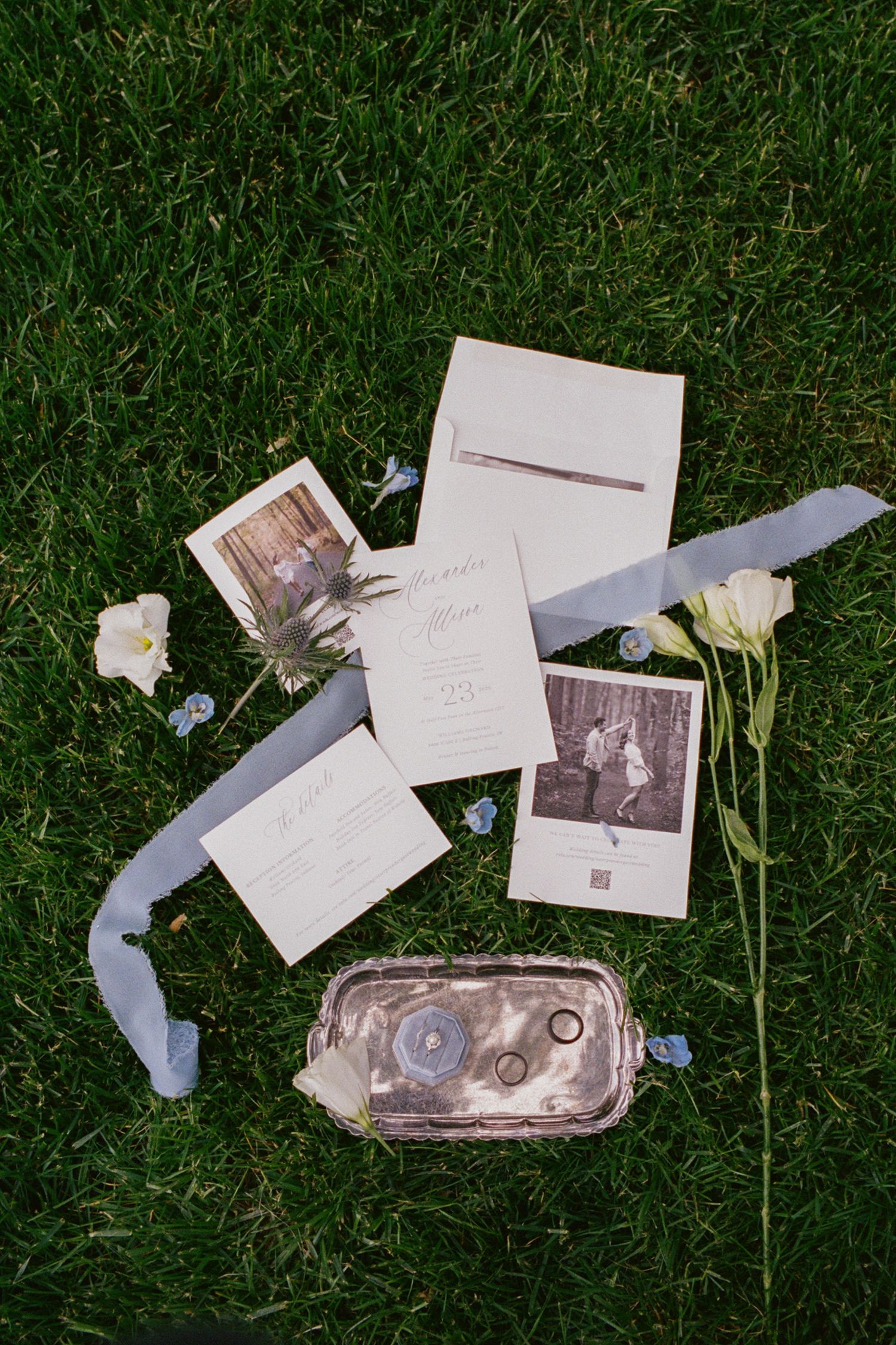 Wedding stationery flat lay on green grass with rings, ribbon, flowers, and vintage photos.