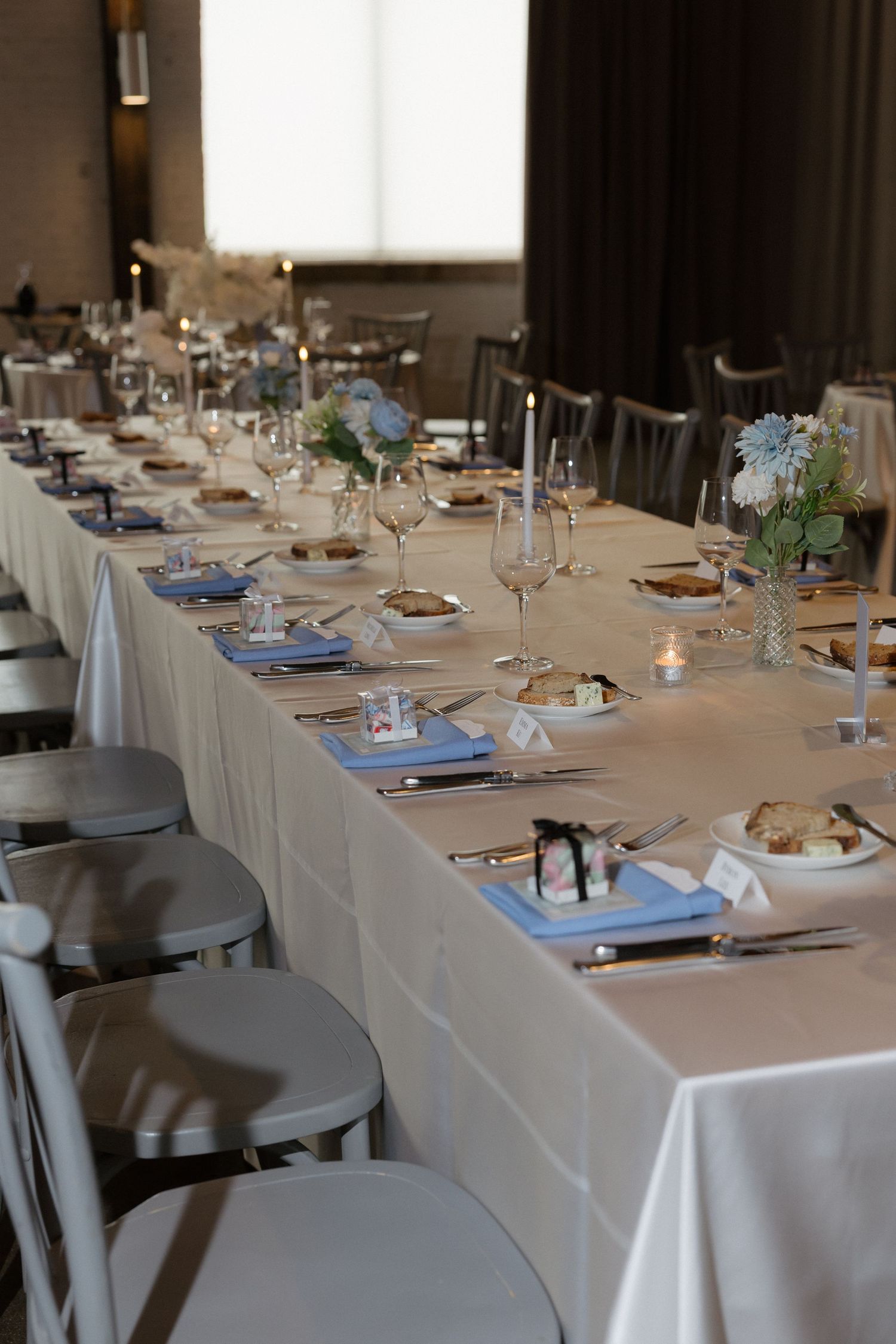 Elegantly set long banquet table with white linen, blue napkins, floral centerpieces, candles, and wine glasses for a formal event.