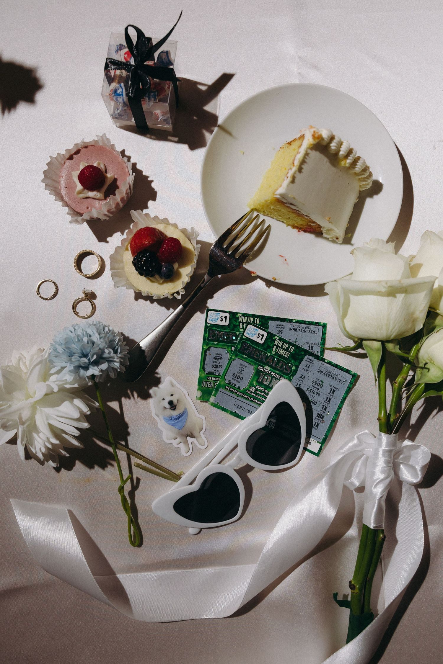 Flat lay of wedding details including cake, cupcakes, rings, lottery tickets, sunglasses, and white roses.