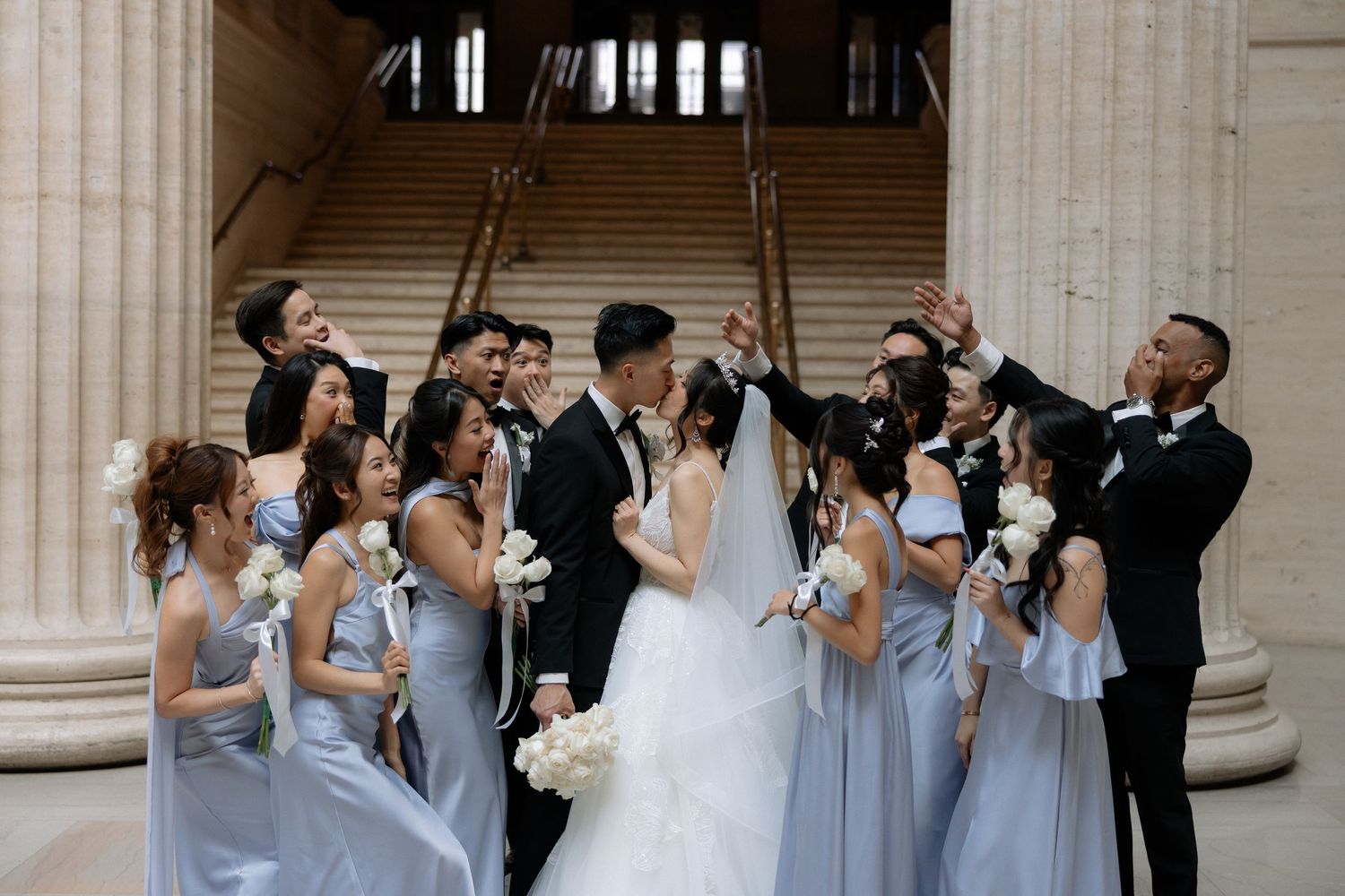 Wedding party celebrates with kiss between bride and groom surrounded by bridesmaids in blue and groomsmen.