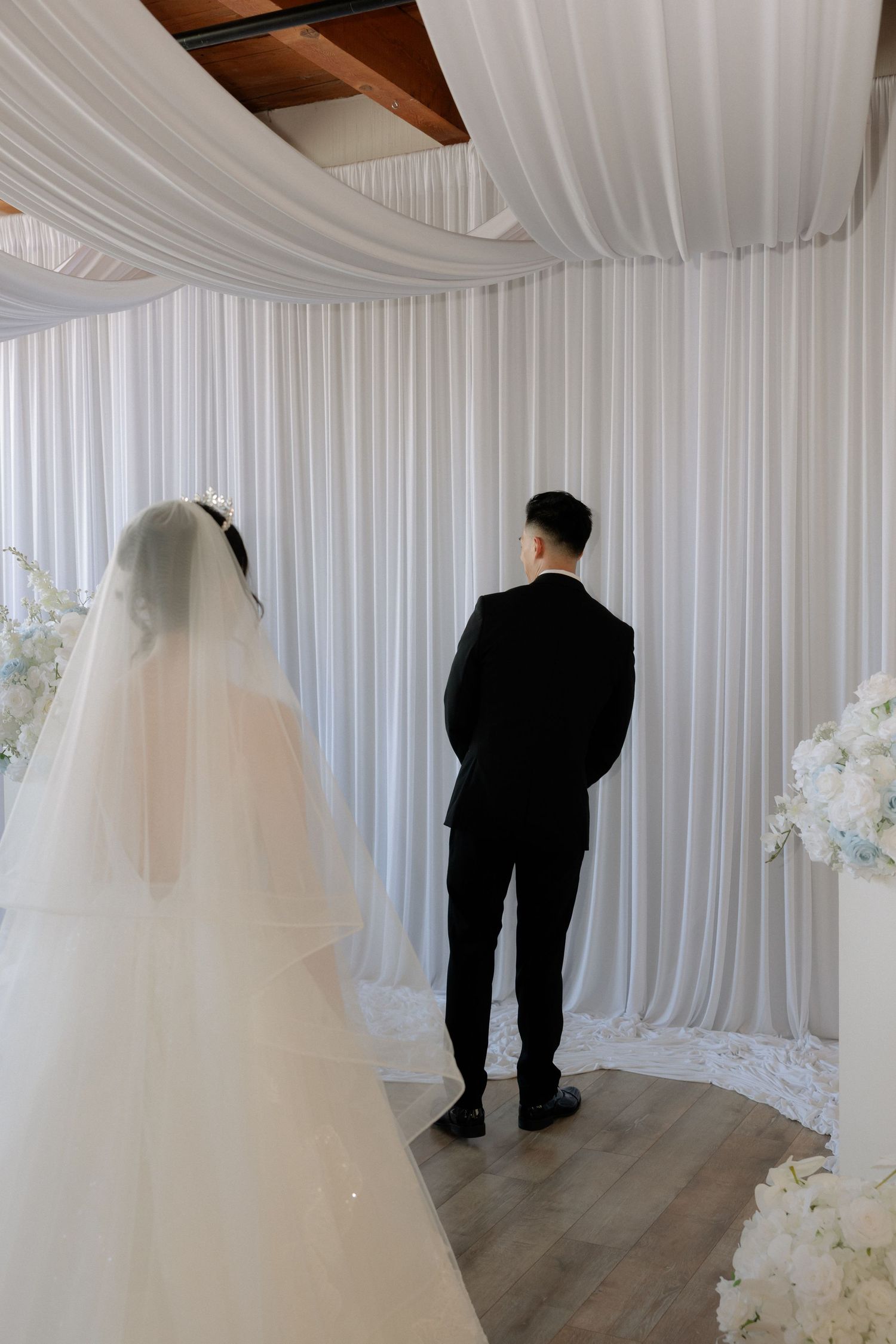 Bride and groom stand before white draped ceremony backdrop in a first look wedding moment.