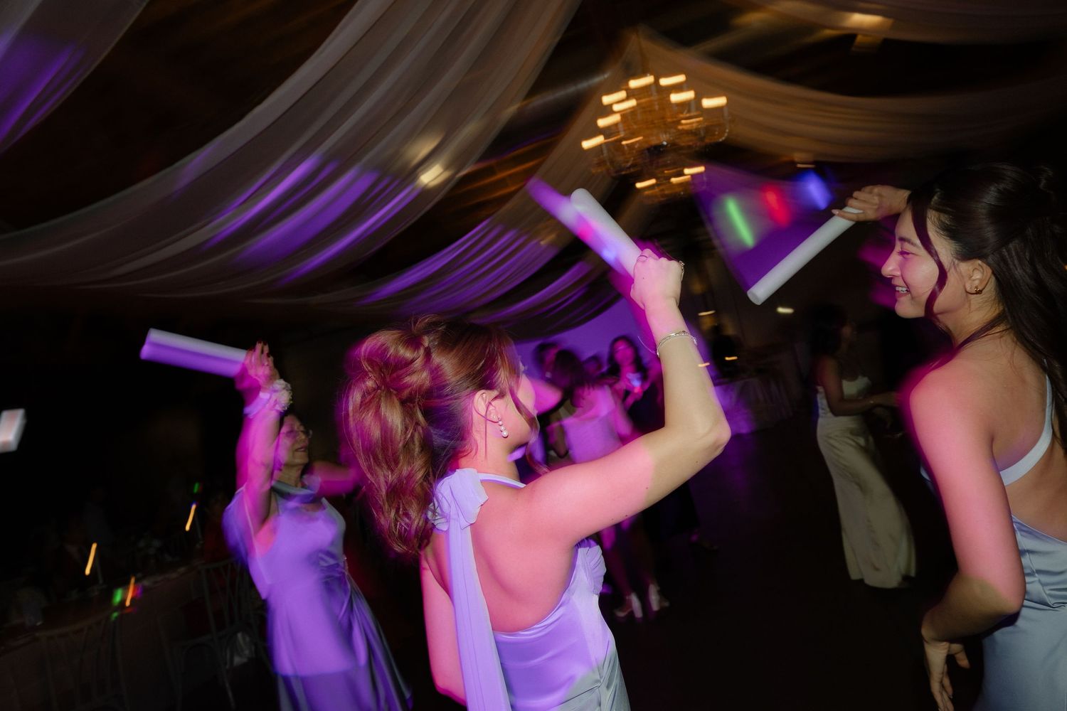 Wedding guests dancing with glowing foam sticks under purple lights on reception dance floor.