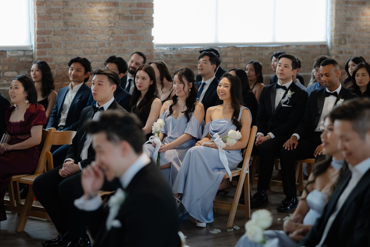 Guests smile and watch a wedding ceremony in a rustic brick venue, bridesmaids in blue dresses seated among attendees.