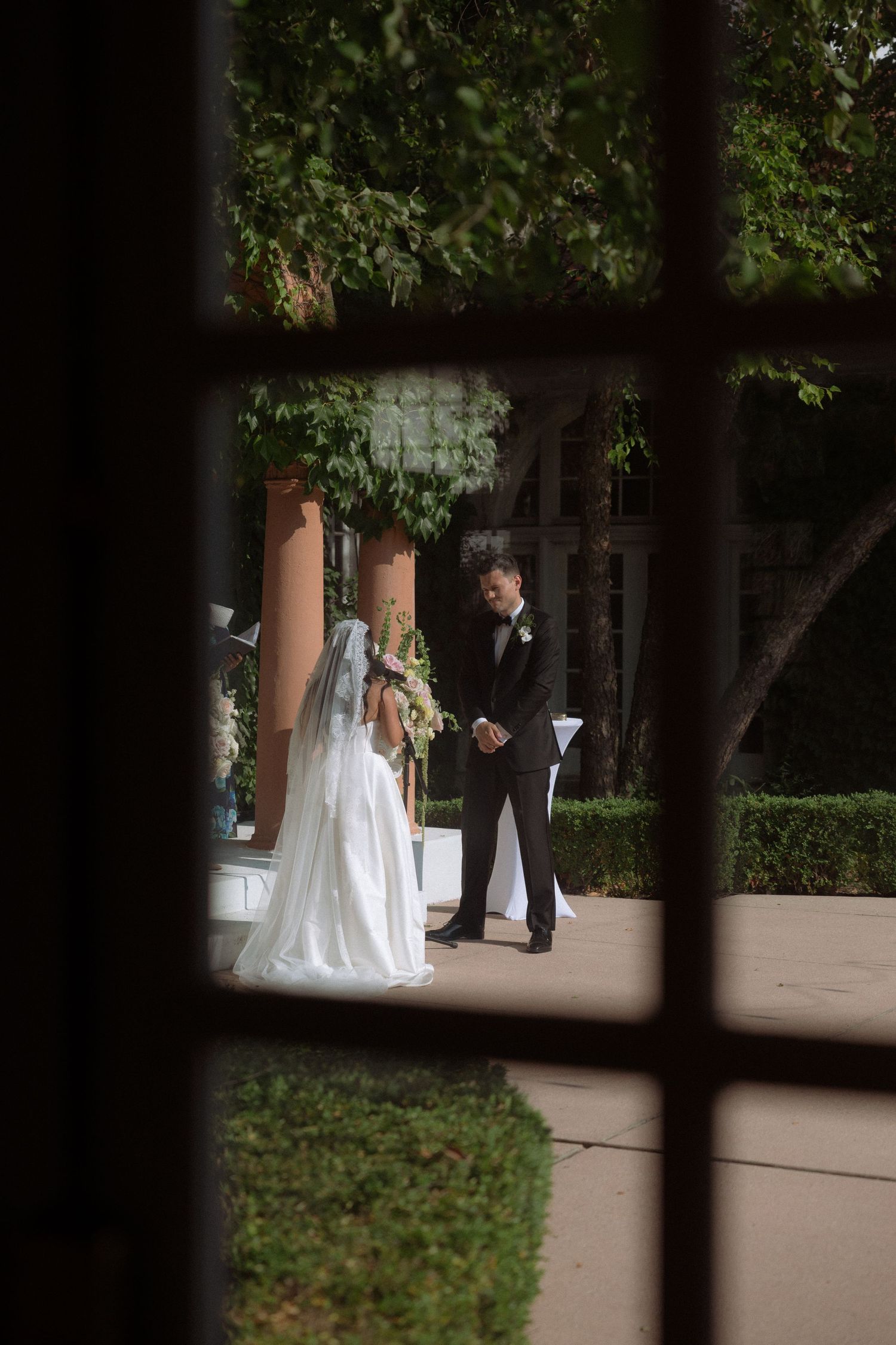 Bride and groom share an intimate moment outdoors, viewed through a window frame.