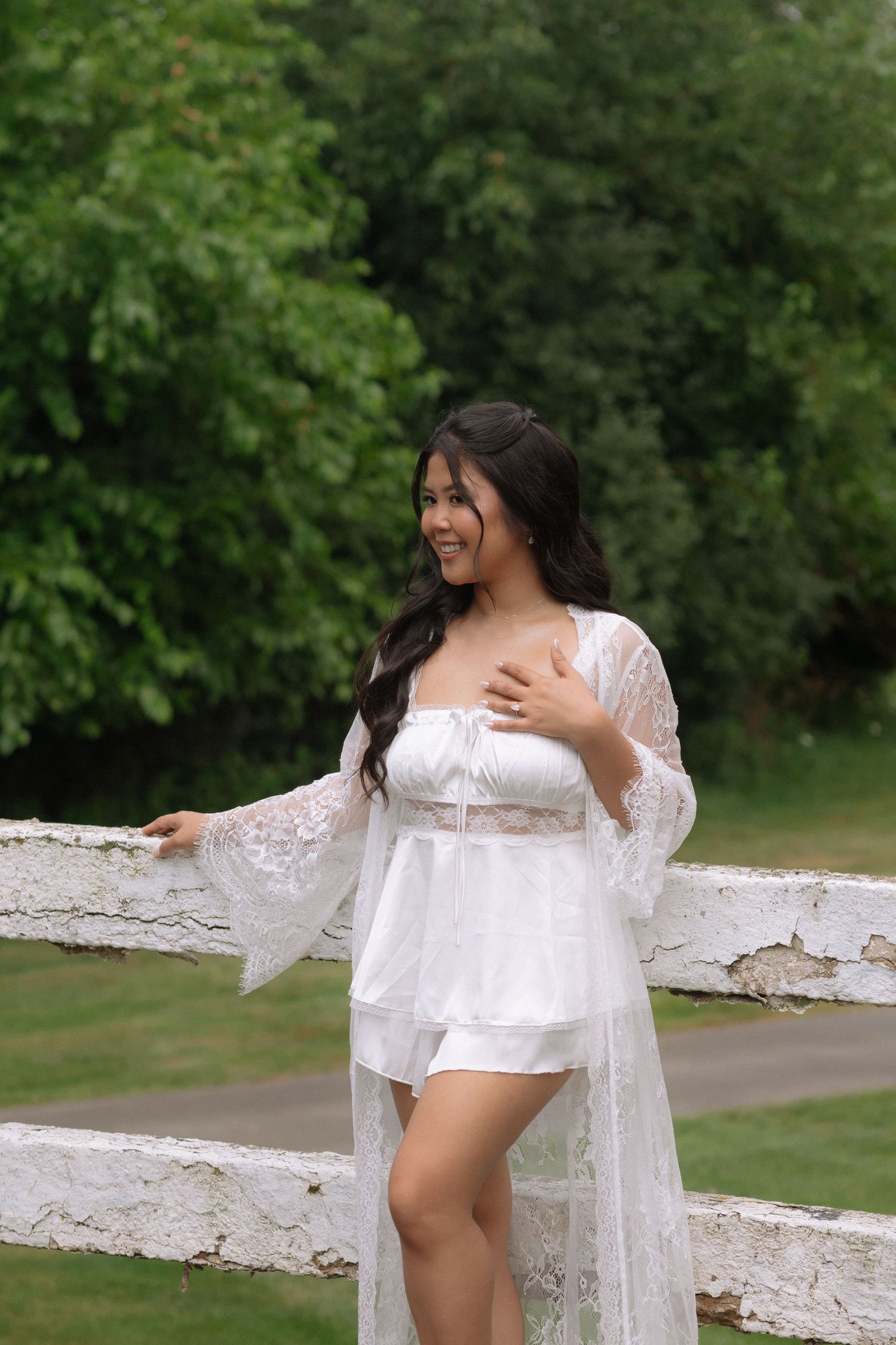 A smiling young woman in white lace lingerie and robe poses gracefully by a rustic white fence in a lush green garden.