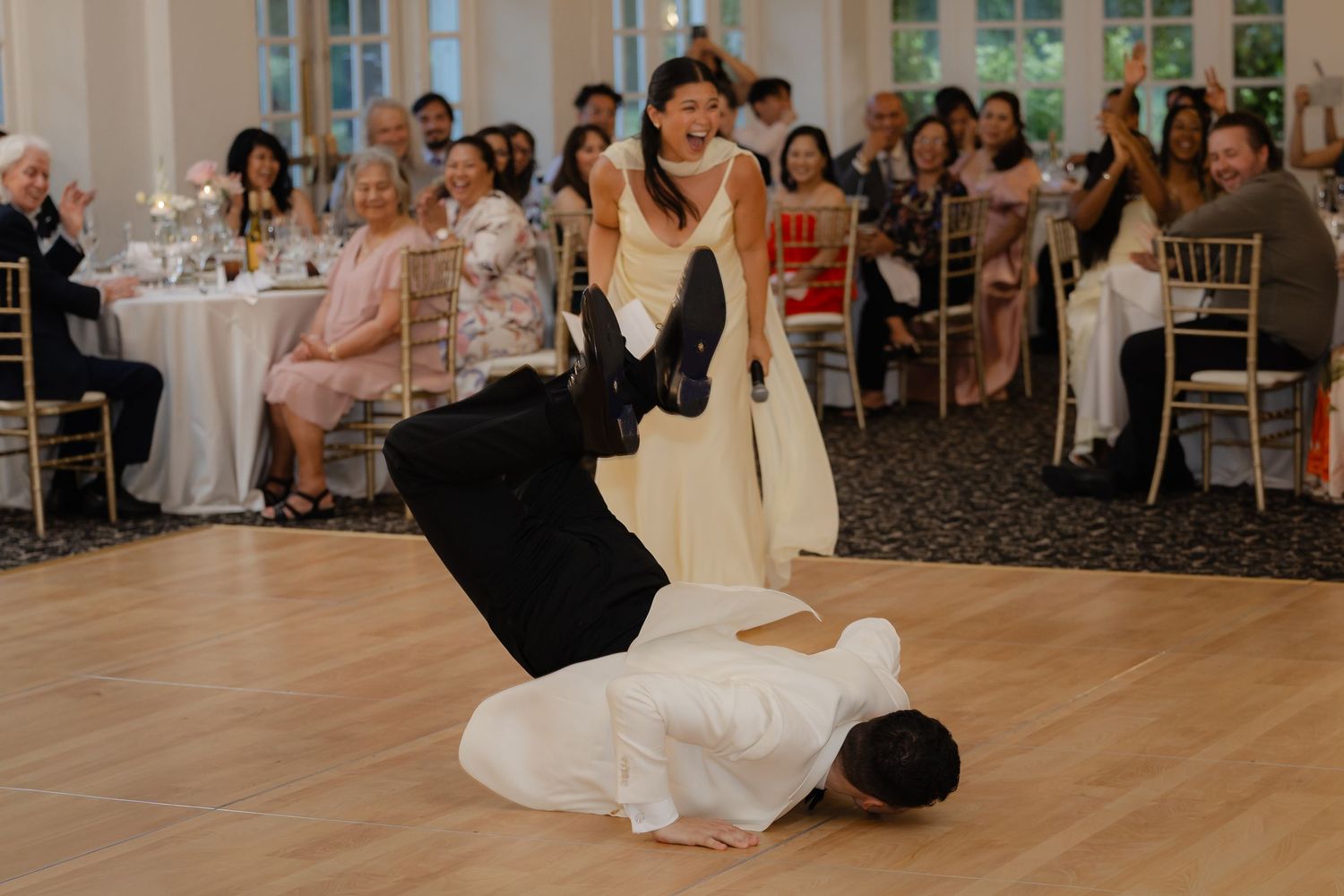Groom breakdances on wedding reception dance floor as bride laughs with guests watching.
