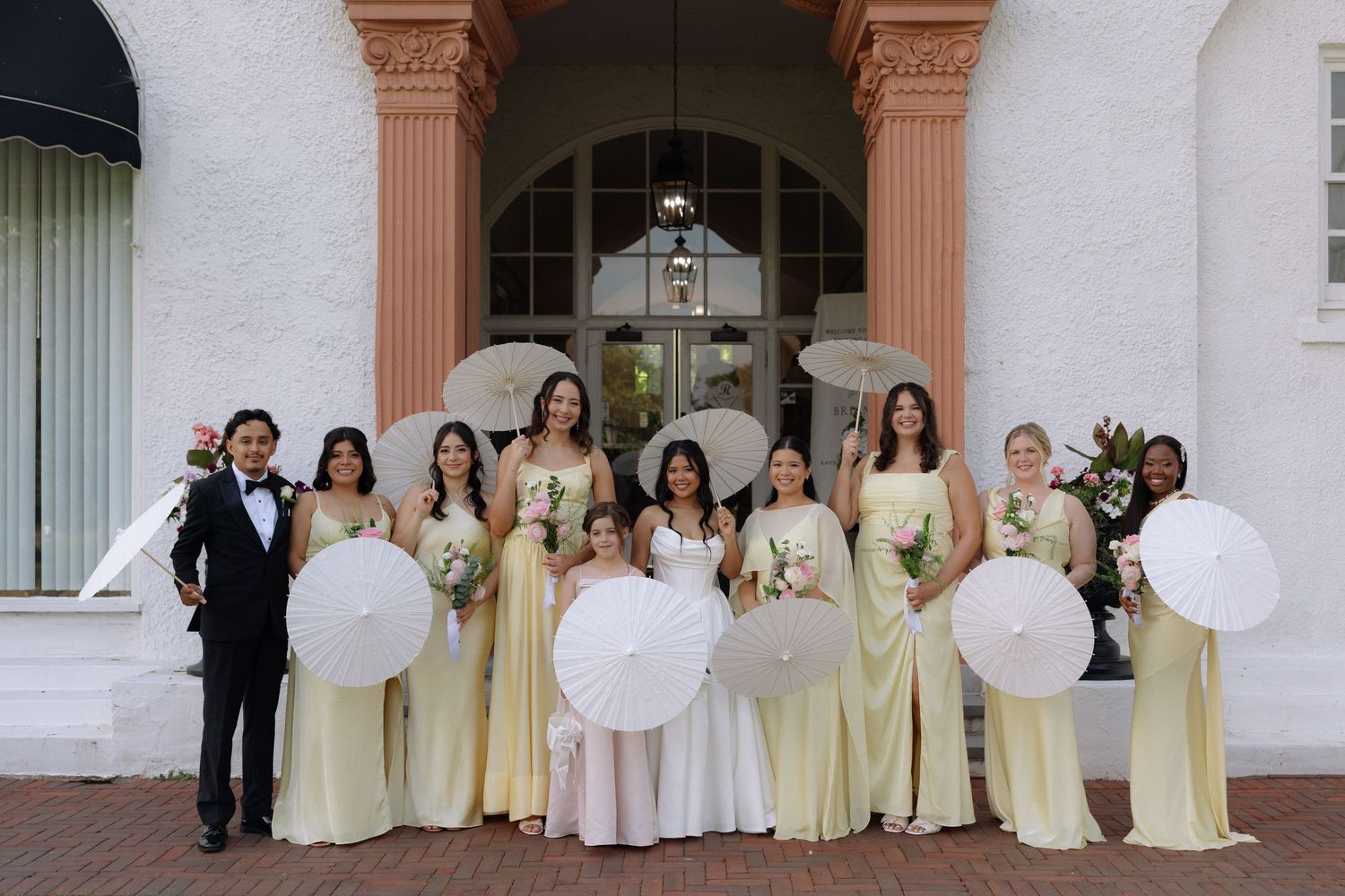 Bridal party in yellow dresses holding white parasols posing in front of elegant white building entrance.