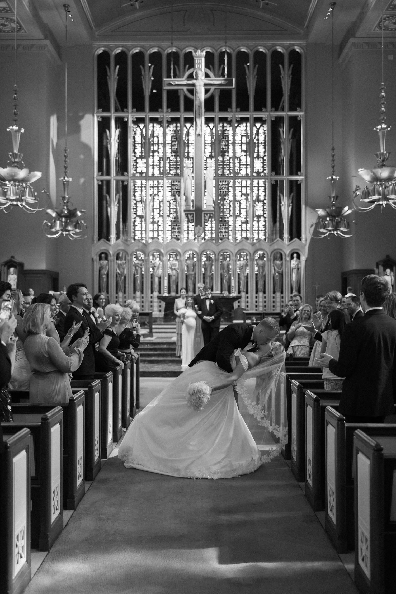 Black and white photo of bride and groom kissing at church altar with stained glass windows and guests applauding.