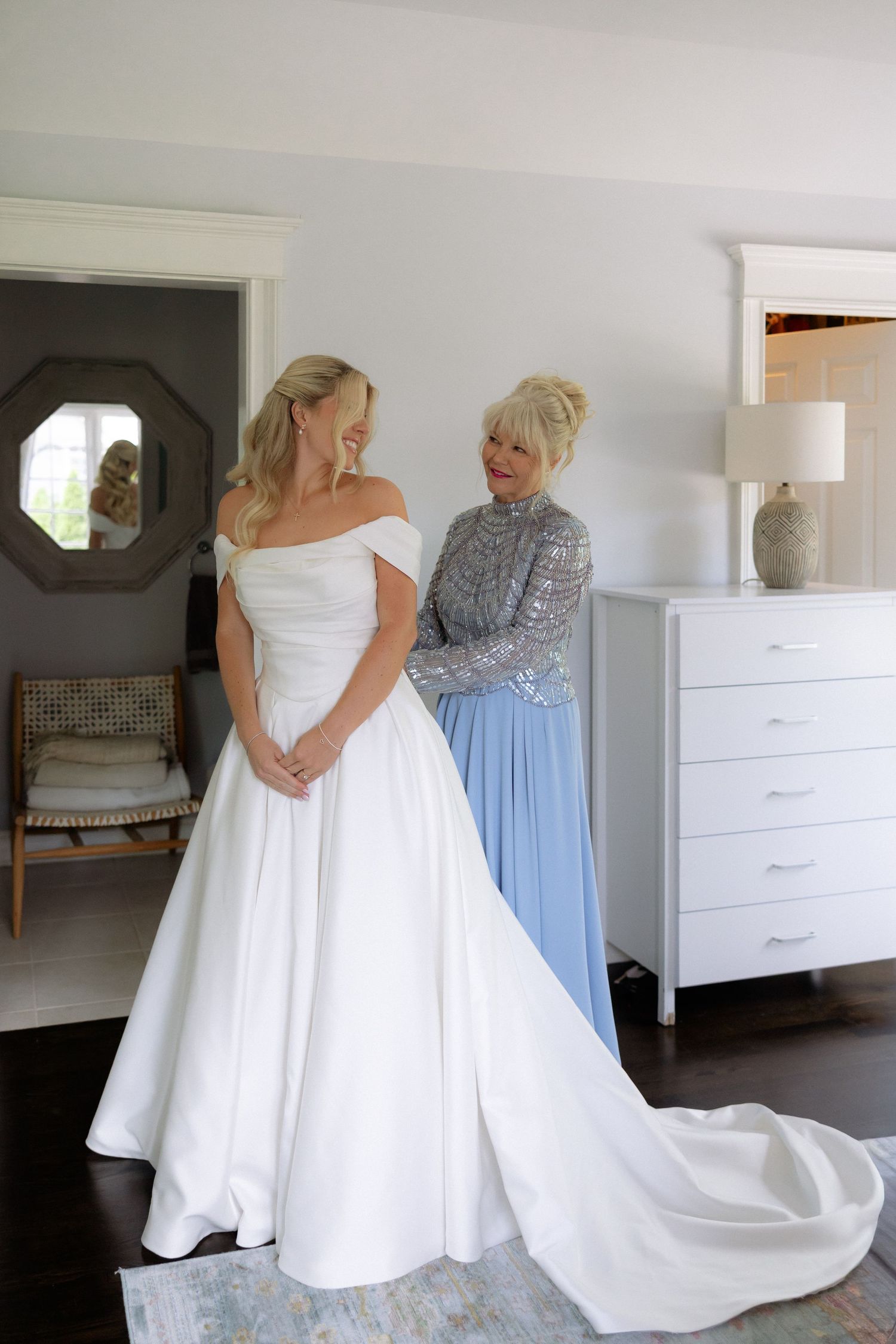 Bride in off-shoulder white gown with train stands with older woman in blue dress in elegant bedroom.
