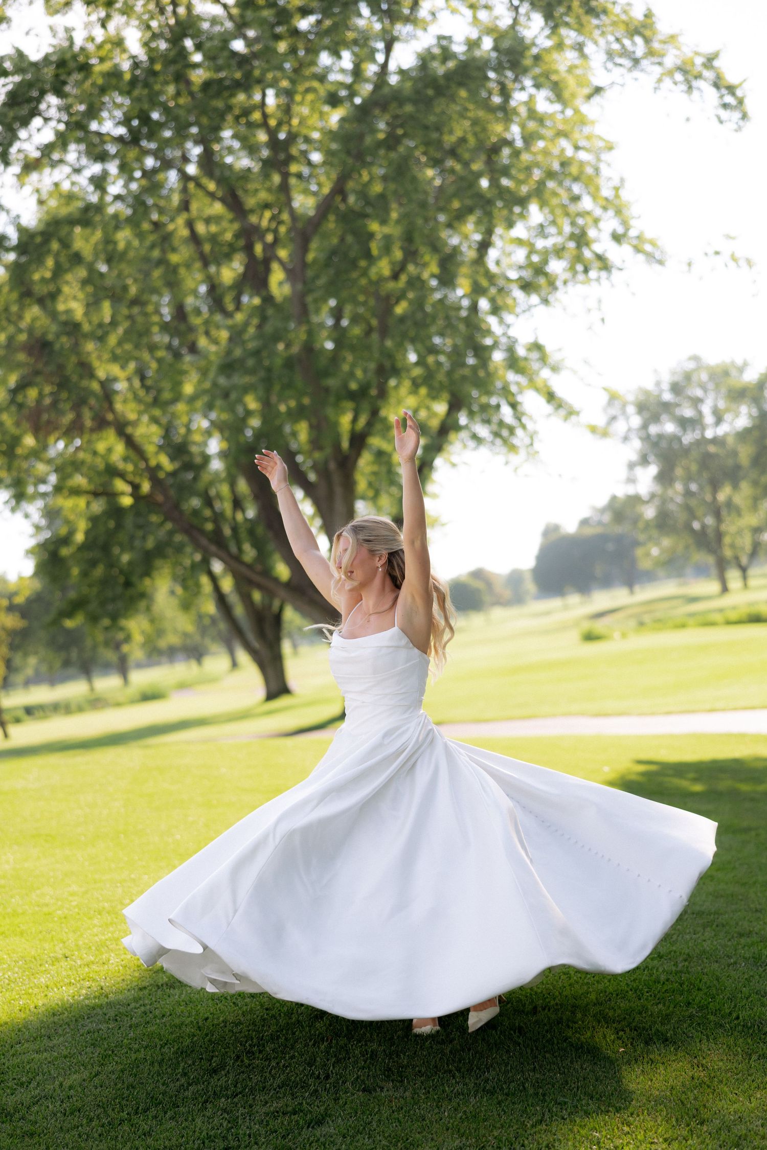 Joyful bride in white satin gown twirling with arms raised on sunny green lawn outdoors.