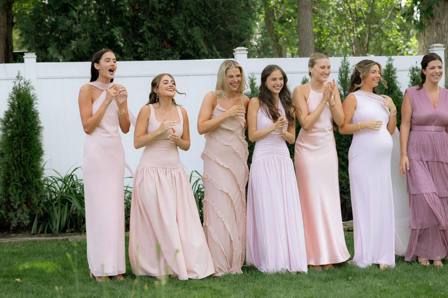 Seven bridesmaids in blush and pink gowns laugh and celebrate outdoors at a summer wedding.
