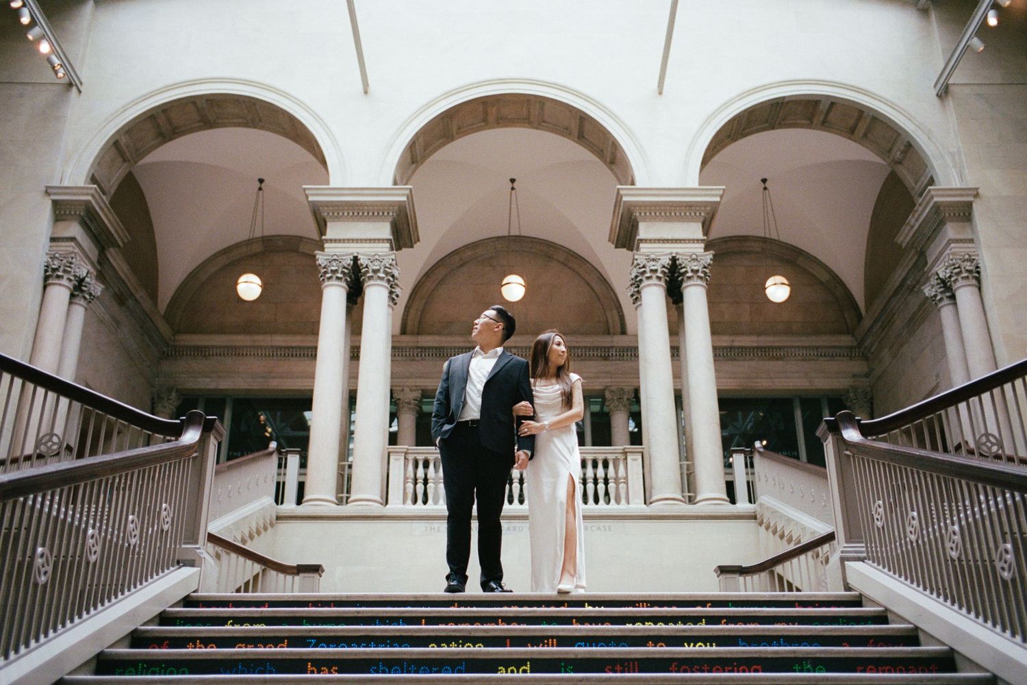 Elegant couple in formal attire standing on grand staircase of a classical building with tall columns.