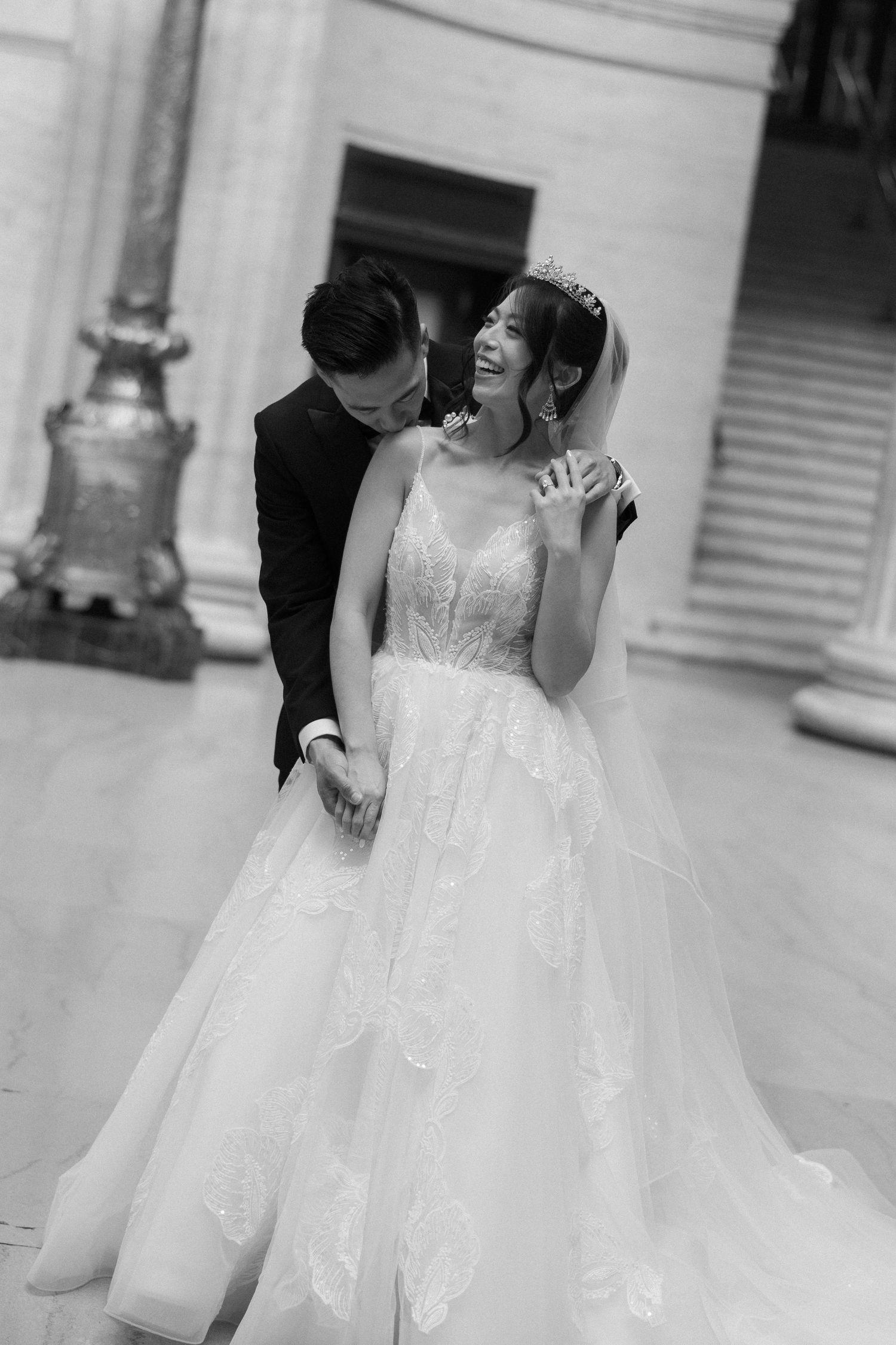 Black and white photo of a smiling bride in a lace ballgown embracing her groom in a classic venue.