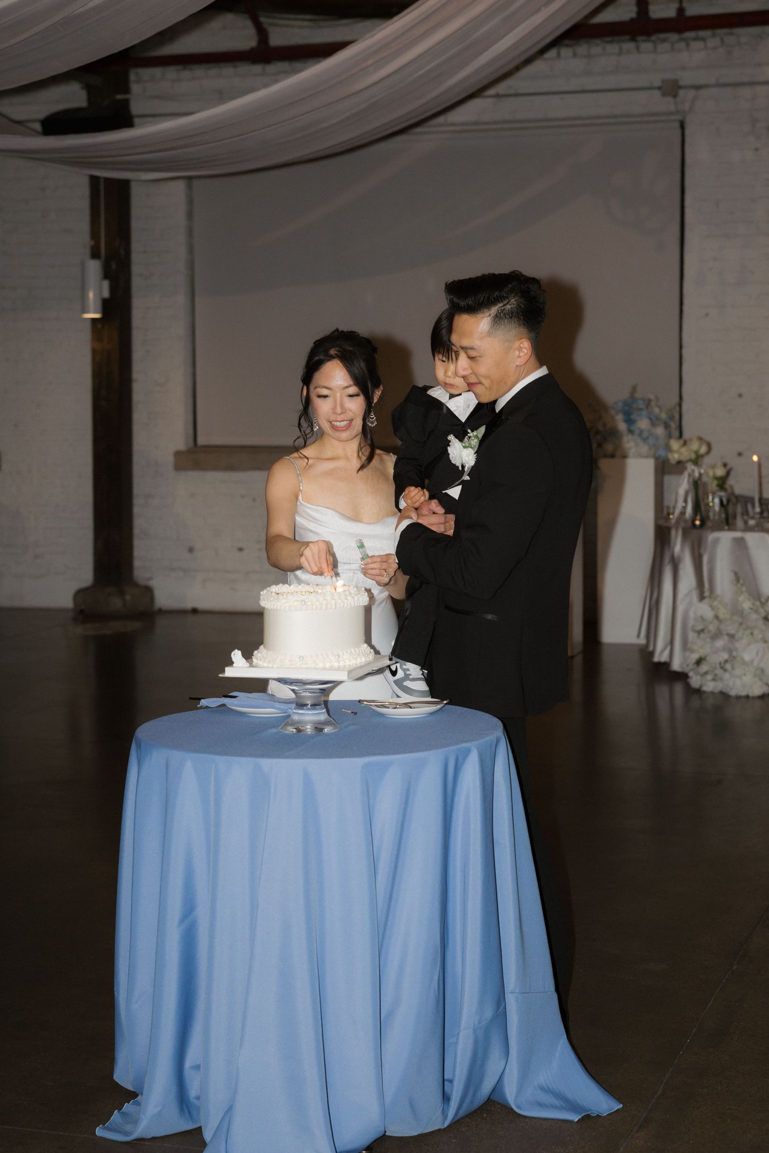 Bride and groom cut white wedding cake together on blue tablecloth at indoor reception venue.