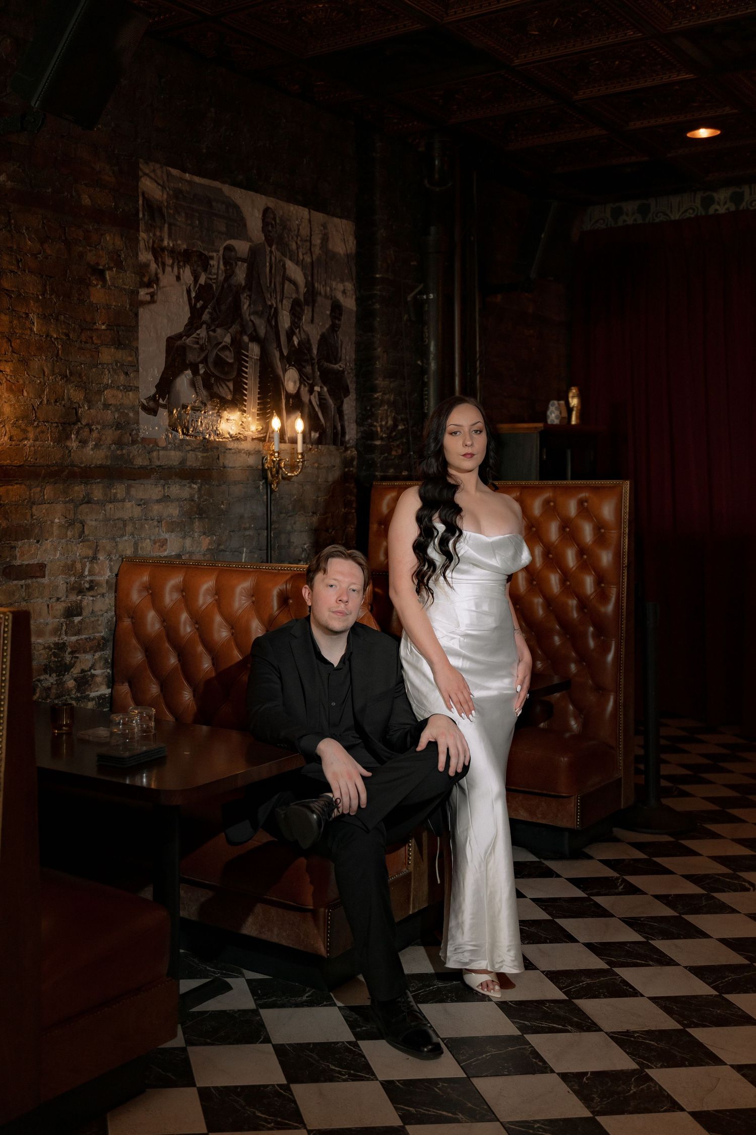 Couple posing in dark elegant bar, man in black suit seated on leather sofa, woman in white satin gown standing beside him.