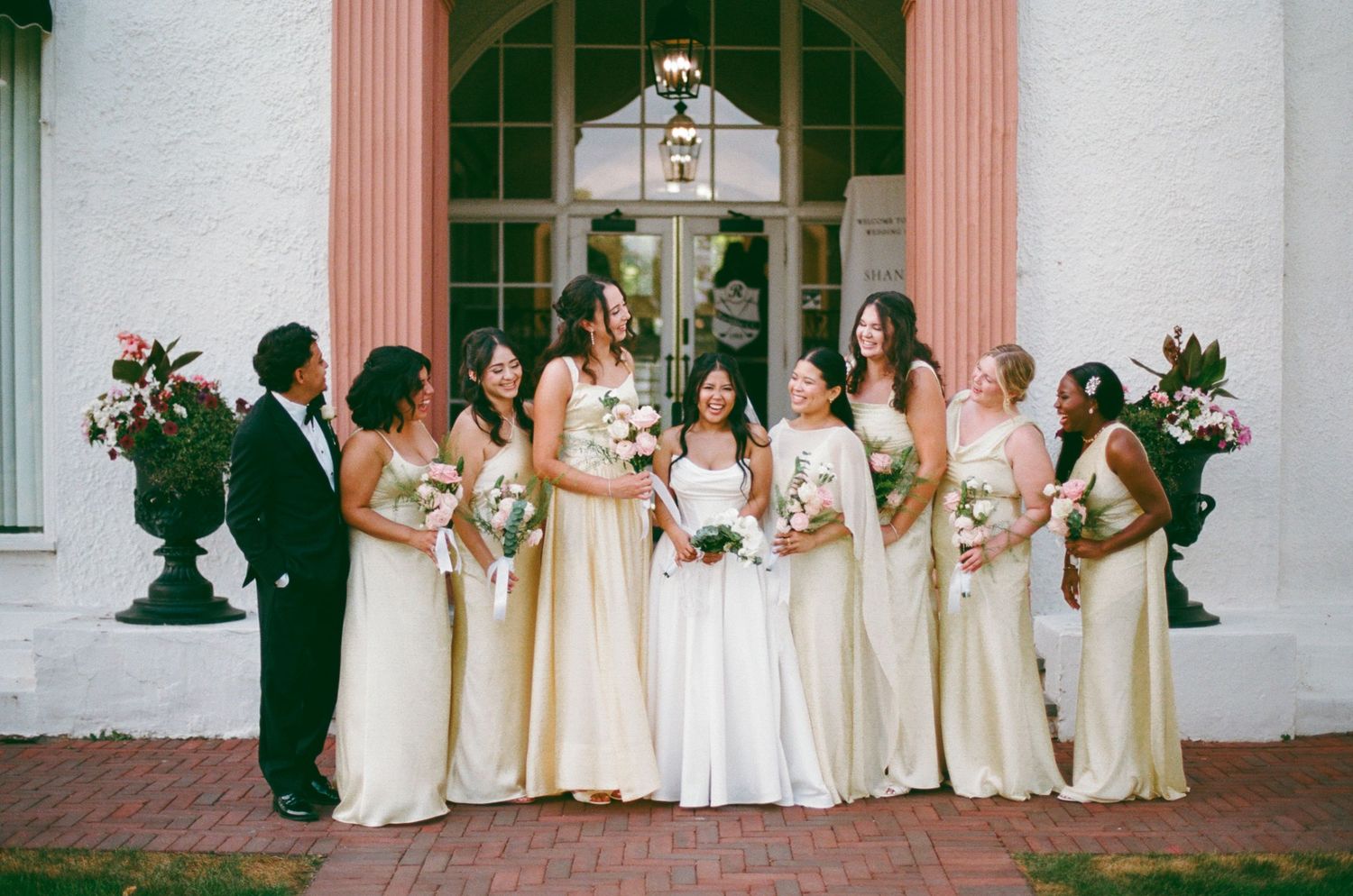 Bride in white gown poses with bridesmaids in champagne dresses holding bouquets outside elegant venue.