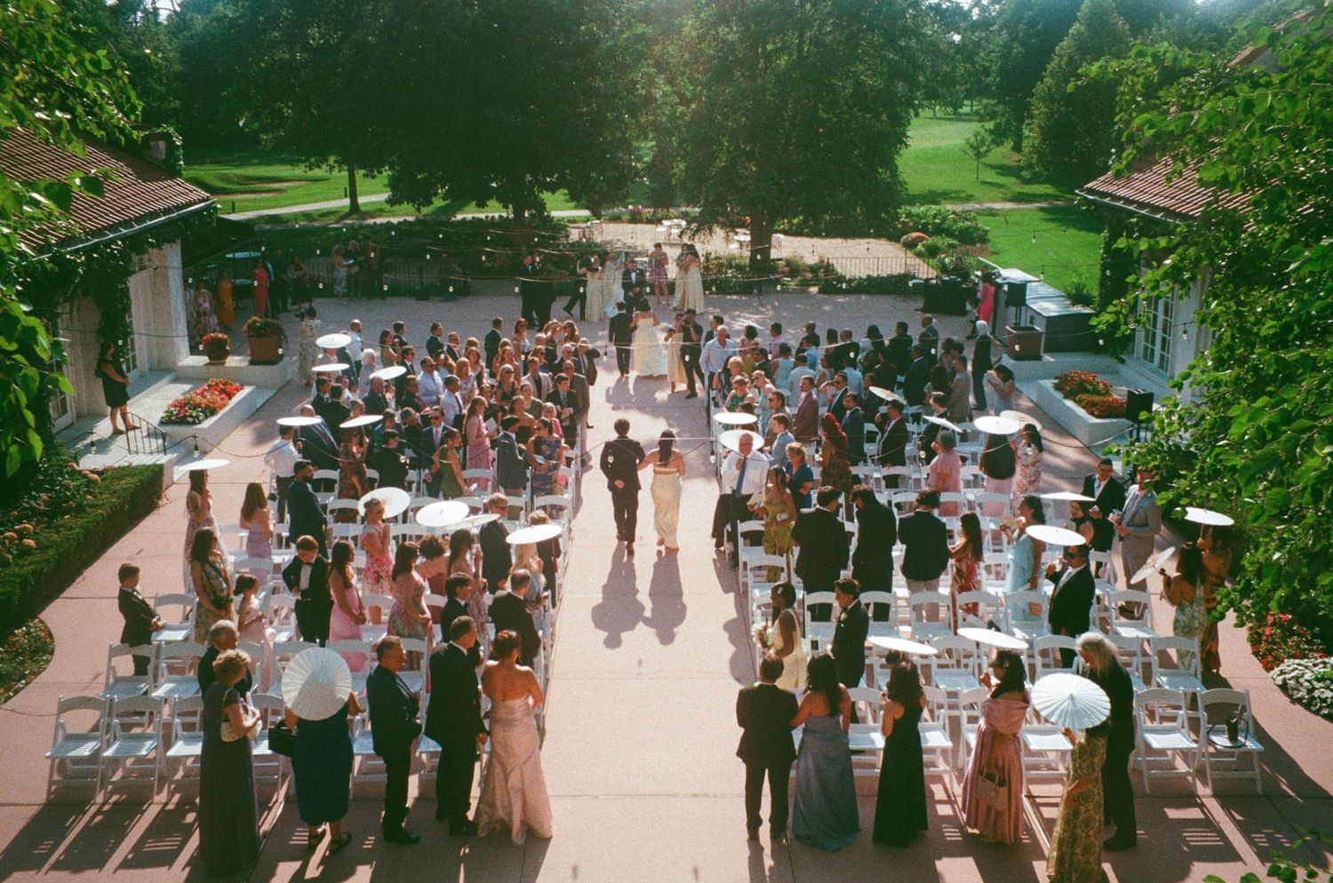 Aerial view of an elegant outdoor wedding ceremony with guests gathered on a brick patio surrounded by lush green trees.