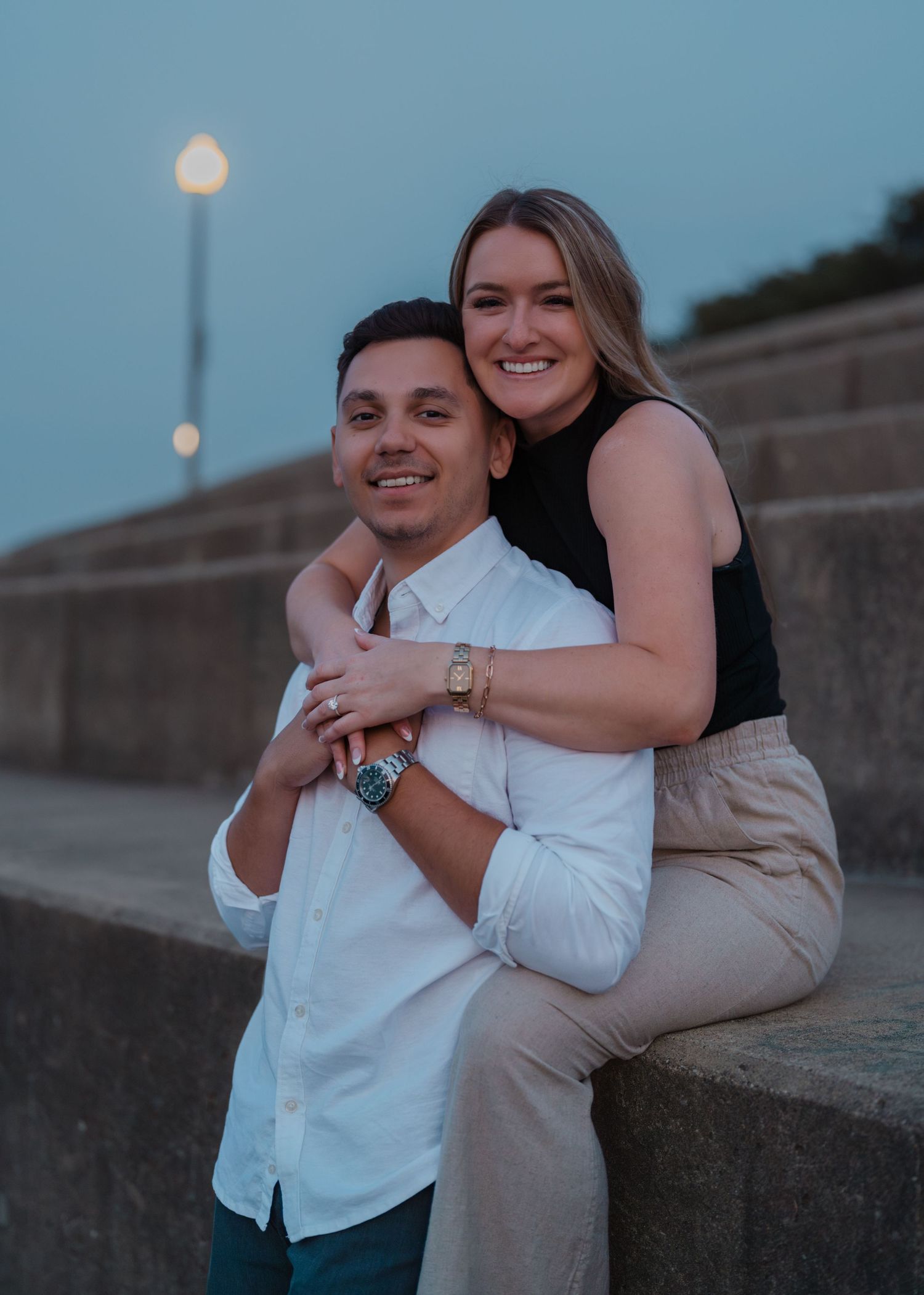 A smiling couple poses at dusk near a stone wall, the woman sitting behind the man with arms wrapped around him.