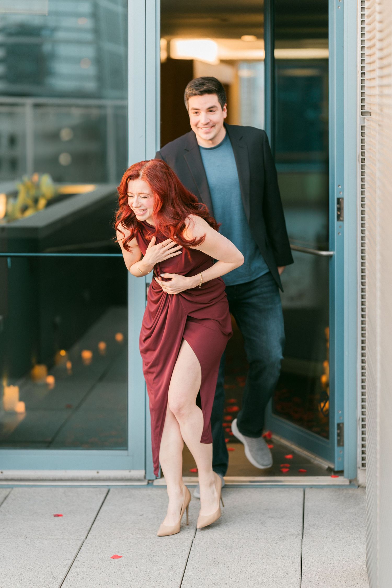 Joyful couple laughing as man embraces woman in burgundy dress stepping outside onto a rooftop terrace.