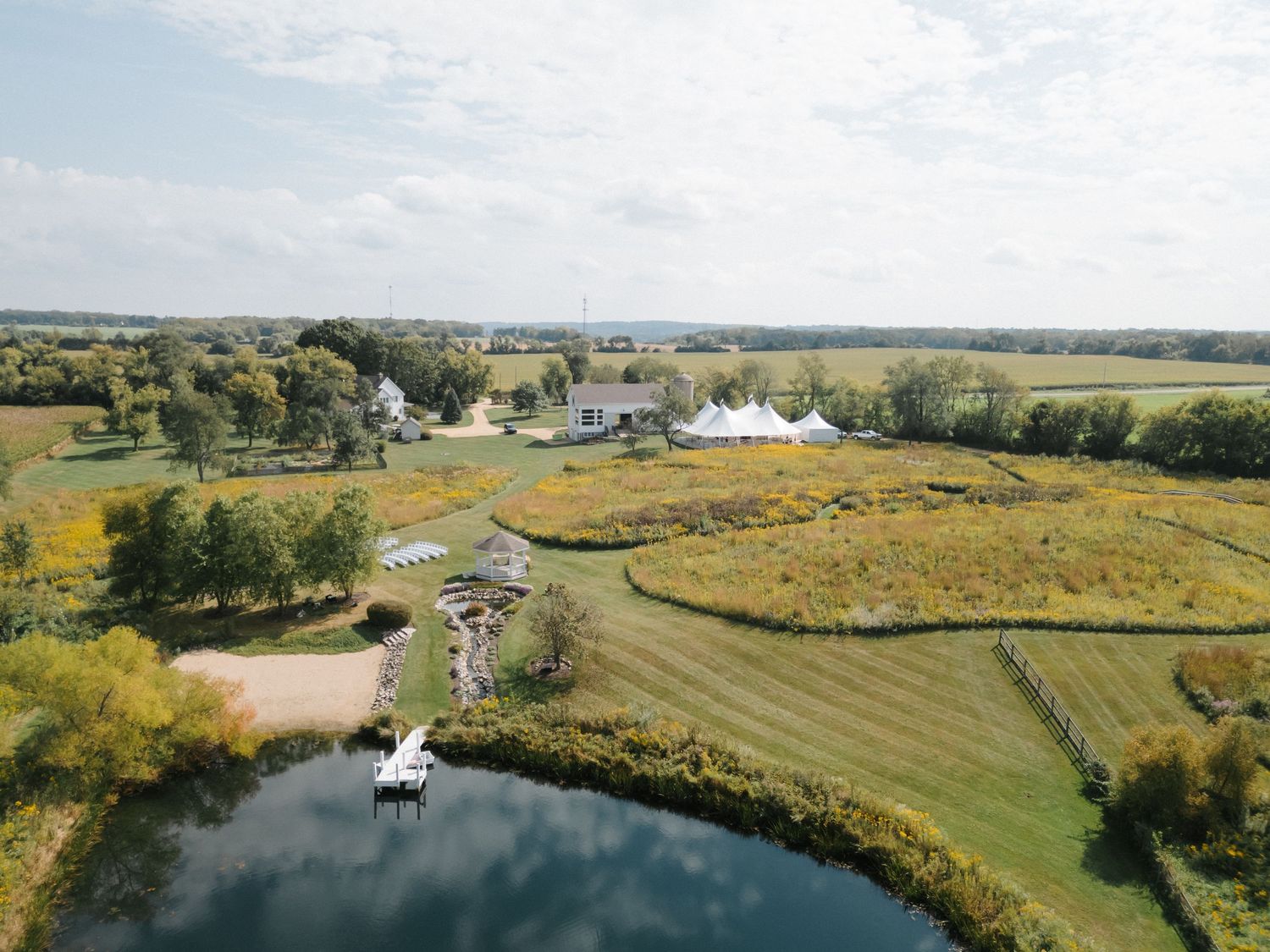 Aerial view of a scenic countryside wedding venue with a pond, white tent, and lush green fields.
