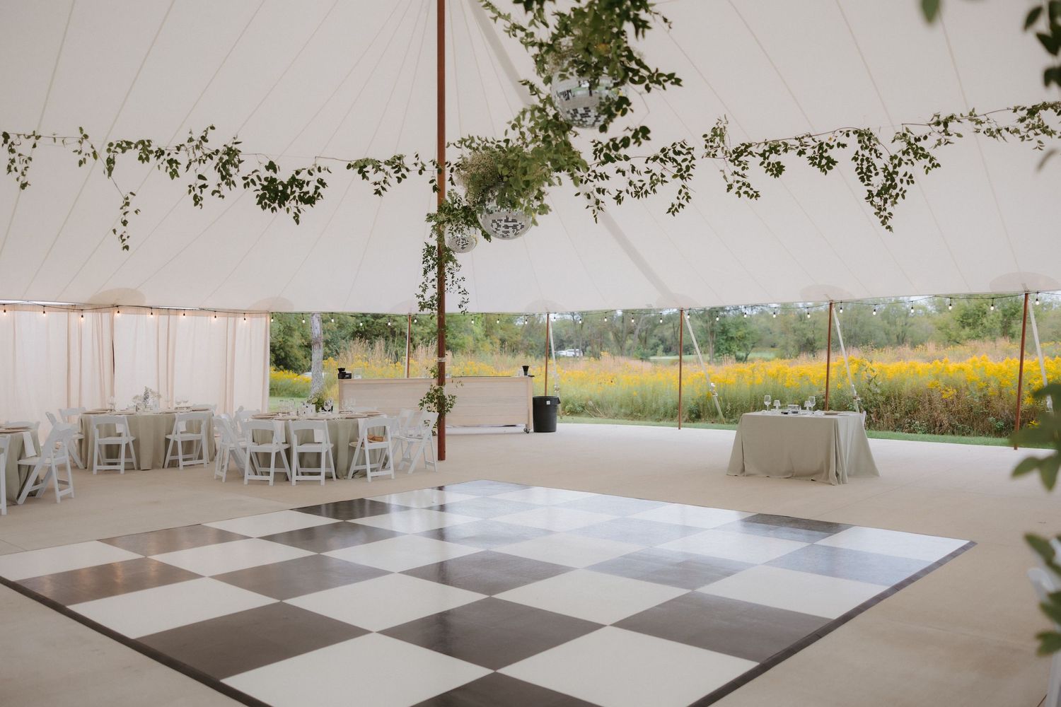 Elegant wedding reception tent with black and white checkered dance floor and yellow wildflower field backdrop.