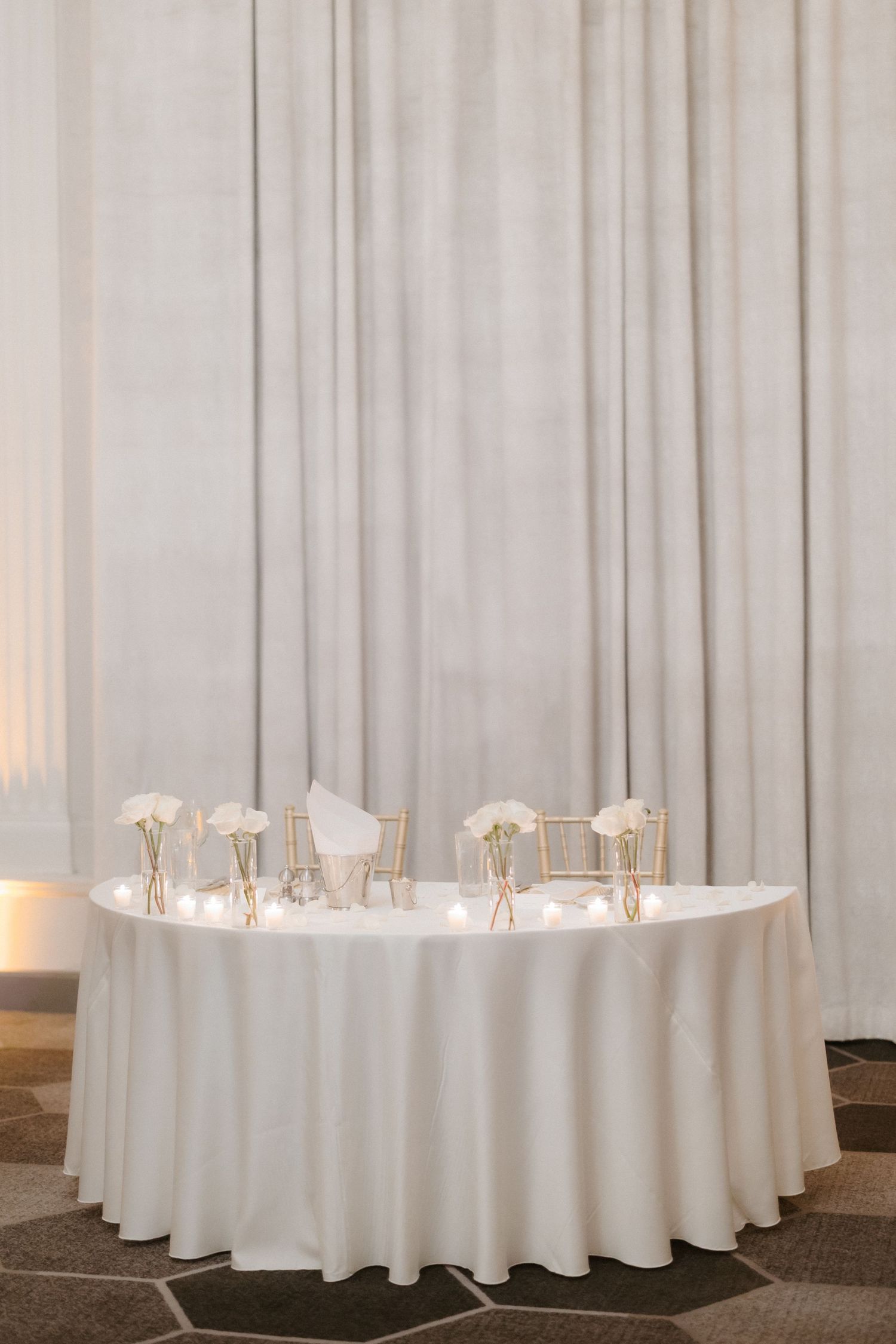 Elegant wedding sweetheart table with white linen, gold accents, and white floral arrangements against draped curtain backdrop.