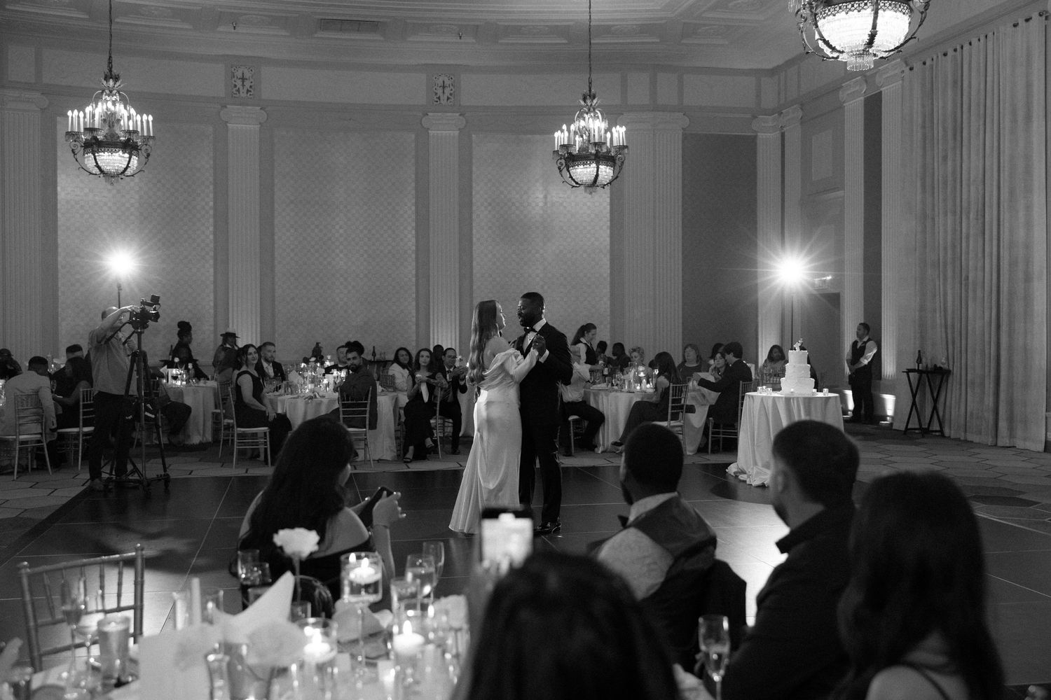 Black and white photo of a couple sharing their first dance at an elegant wedding reception with chandeliers.
