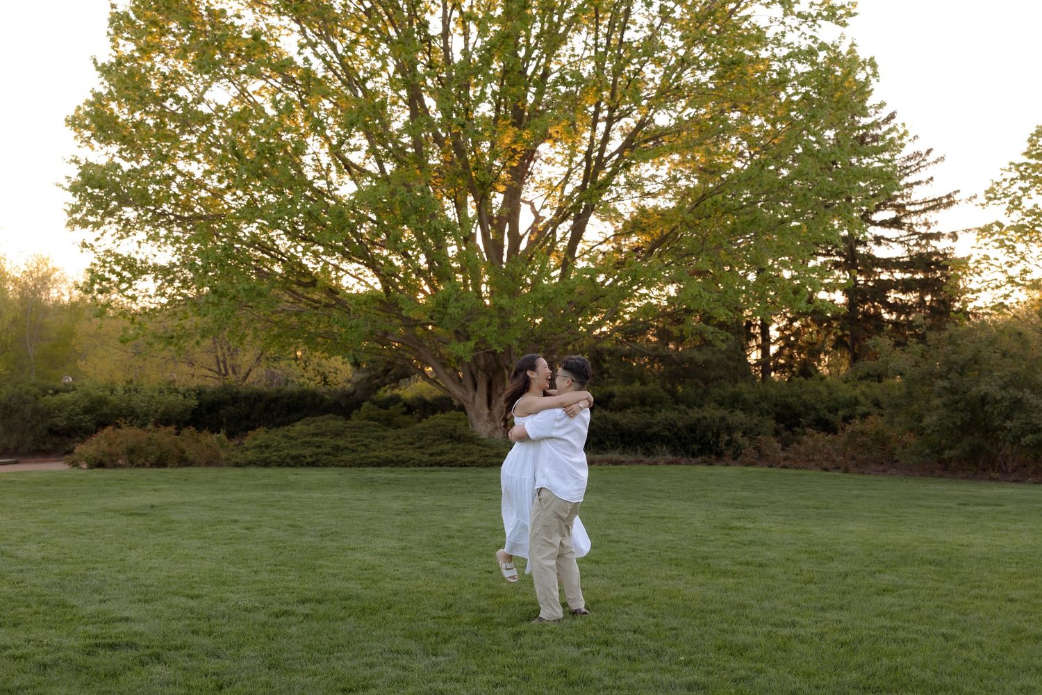 Couple embracing and spinning in lush green garden at golden hour with large tree backdrop.