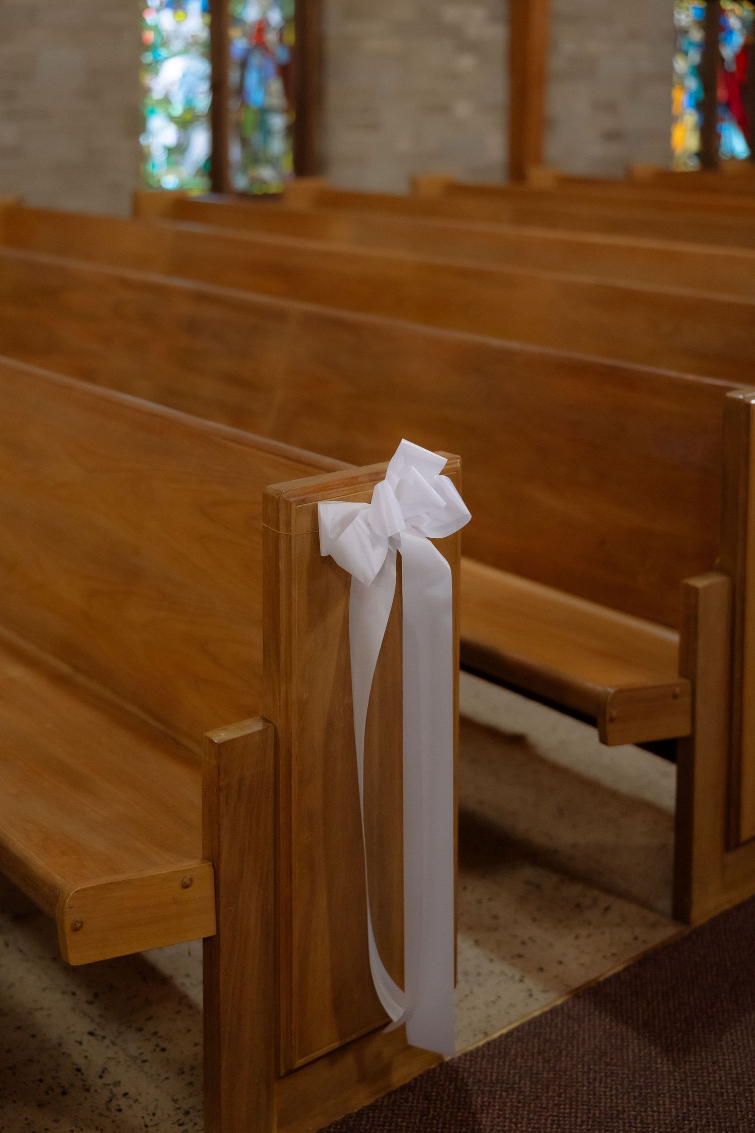 Wooden church pews decorated with white ribbon bows for a wedding ceremony, with stained glass windows in background.