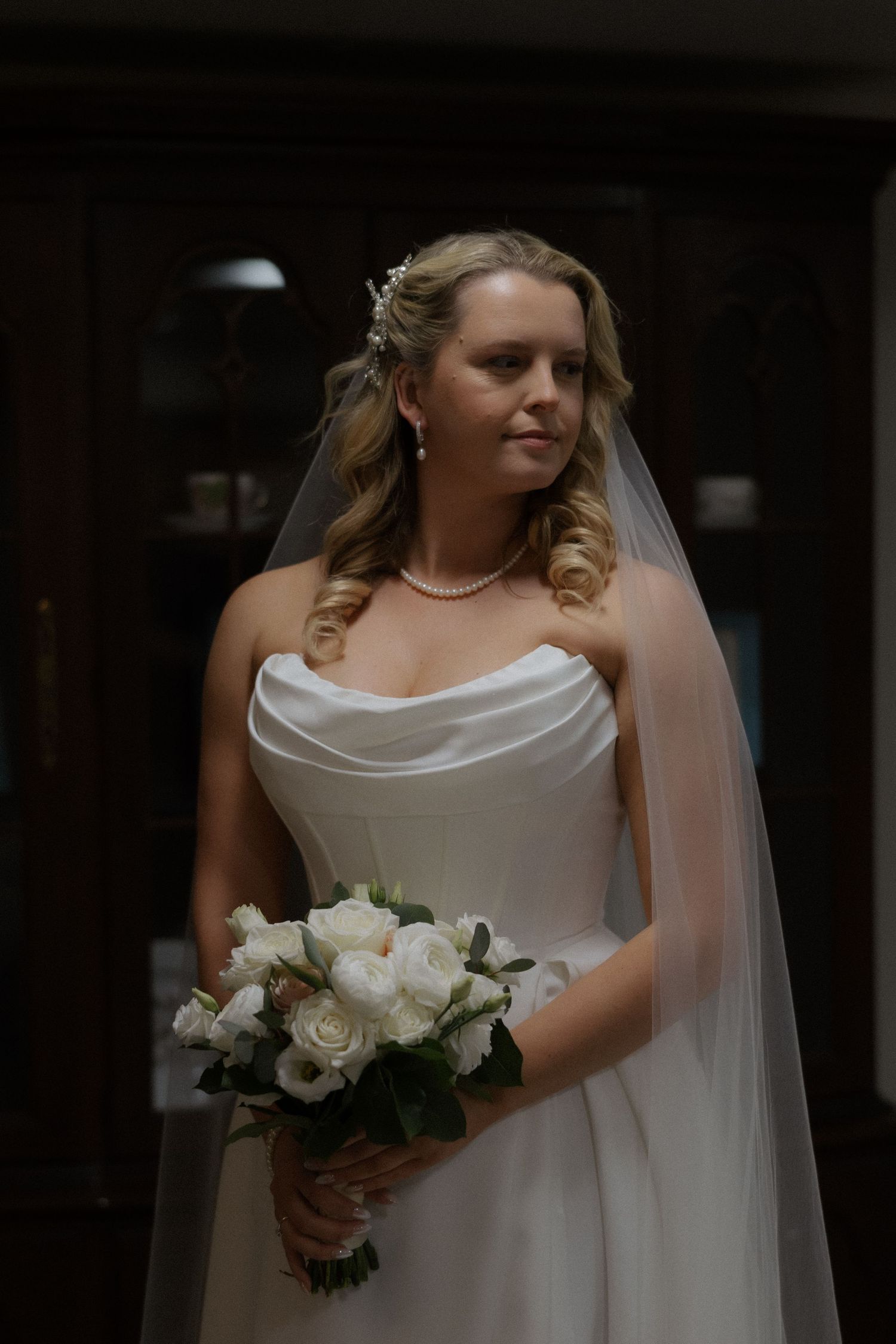Elegant bride in strapless white gown holding bouquet of white roses, wearing pearl necklace and veil.
