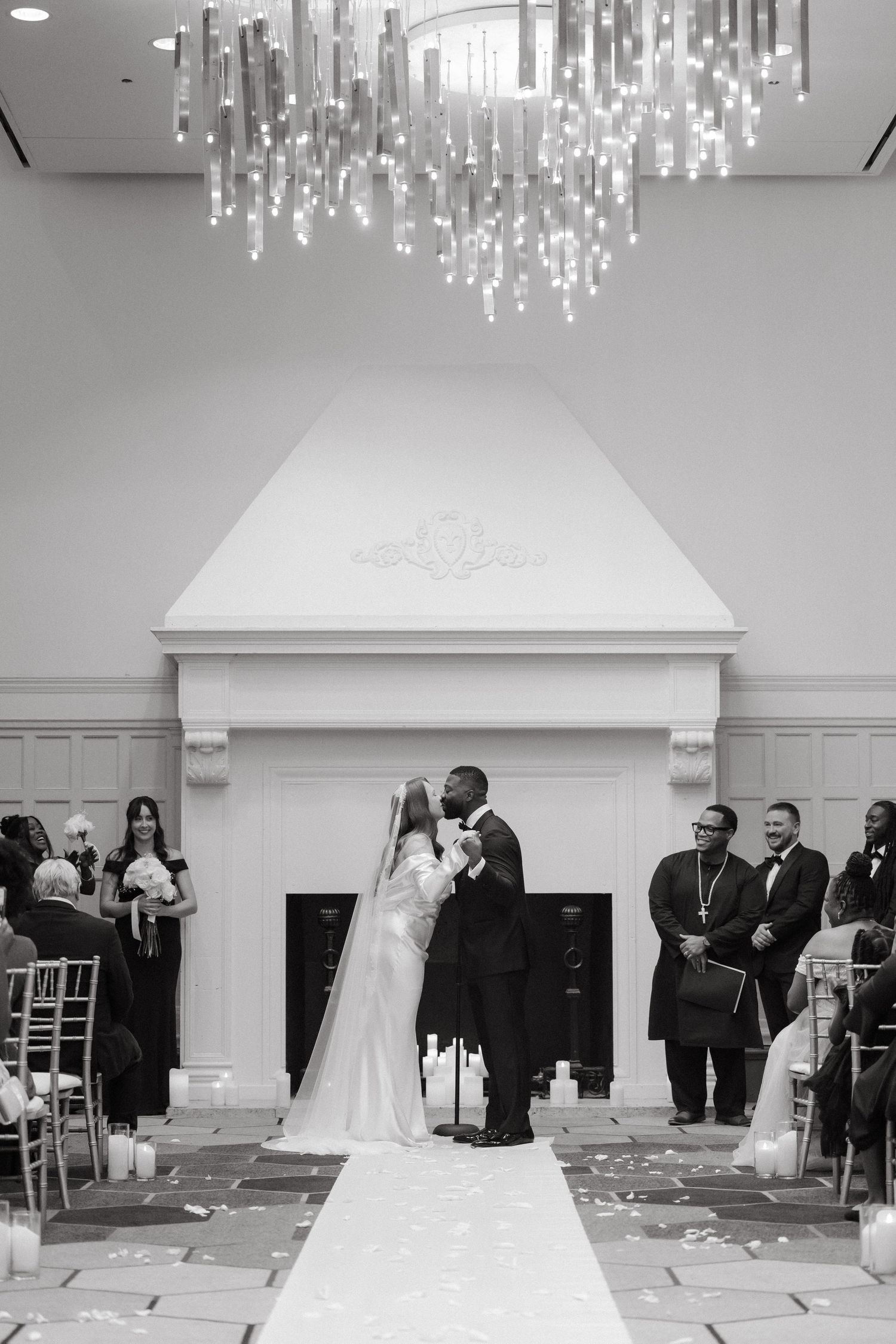 Black and white wedding photo of couple sharing first kiss at altar under elegant chandelier in grand ballroom ceremony.