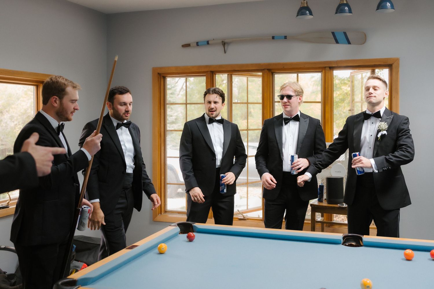 Five groomsmen in matching black tuxedos socializing around pool table before wedding.