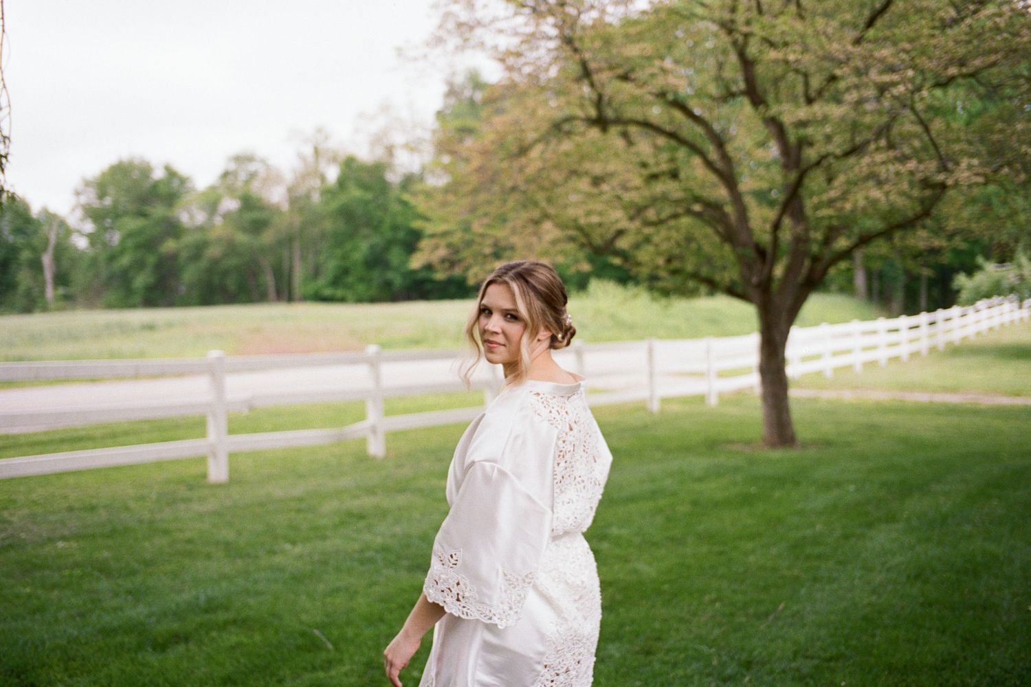 A woman in a white lace dress walks gracefully on a green lawn with a white fence and trees in the background.