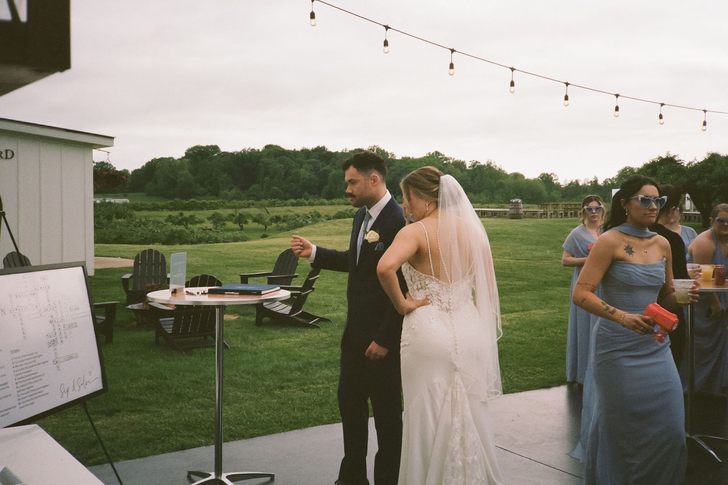 Bride and groom review seating chart at outdoor wedding reception with string lights overhead.