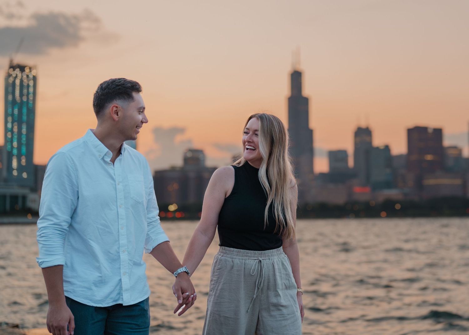 Couple holding hands smiling at sunset with Chicago skyline and Willis Tower in background near lakefront.